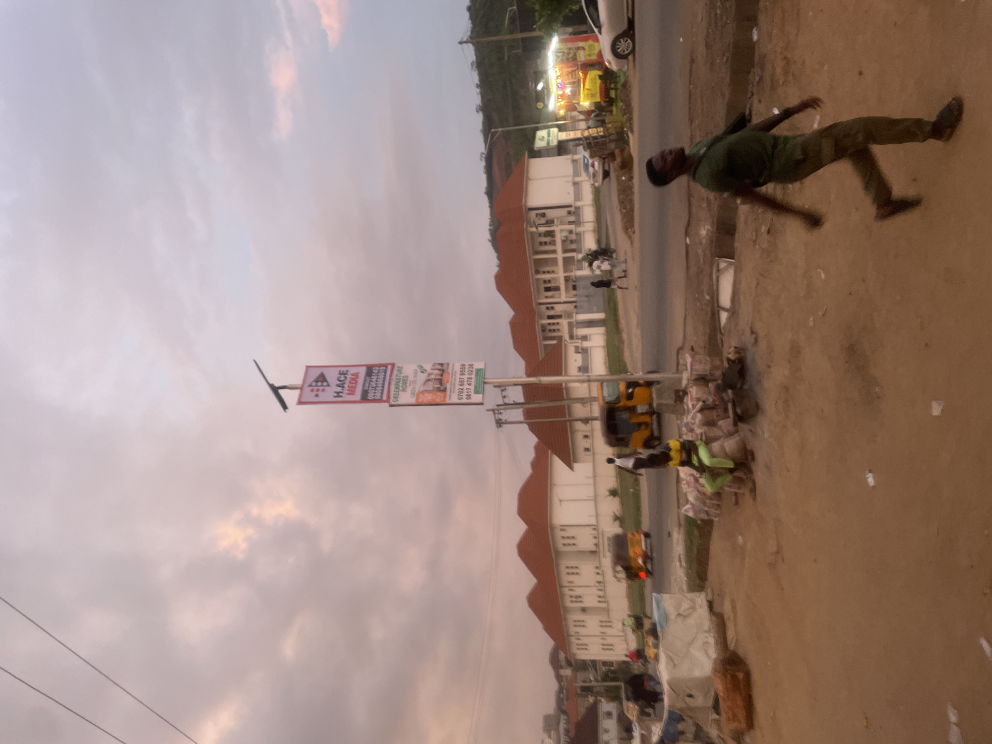 The image shows a street scene in Abuja, Nigeria. A man in dark green clothing is walking on the right side of the frame. Beside him is a dirt area. Adjacent to this space there is a yellow and green tricycle. A road runs parallel to the man and tricycle. On the other side of the road, there are white apartment buildings with red tiled roofs. A large sign with "H. ACE MEDIA" is visible. The sky is overcast with light pink and grey tones.