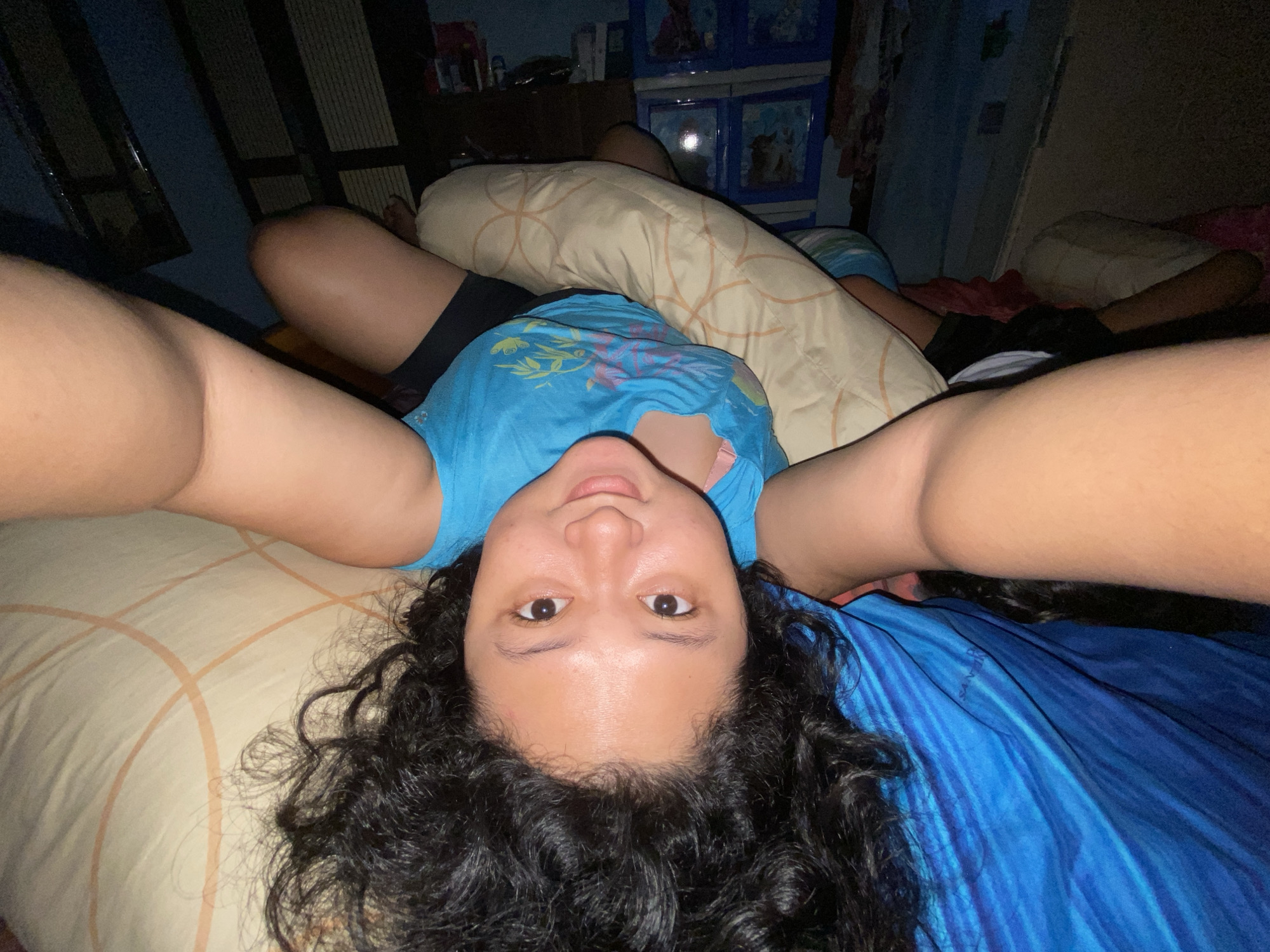 A person is lying on their back on a bed, with their head at the bottom of the frame and their feet at the top. The person is wearing a blue t-shirt with a graphic print. Their legs are spread apart, and their arms are extended upwards. The bed is covered with a light-colored comforter with a geometric pattern and a body pillow with a similar pattern. In the background, there is a dark wooden dresser and a blue plastic cabinet with drawers. The scene appears to be indoors, likely a bedroom. The lighting is dim. There is no indication of the person's location in Gelam, Indonesia.