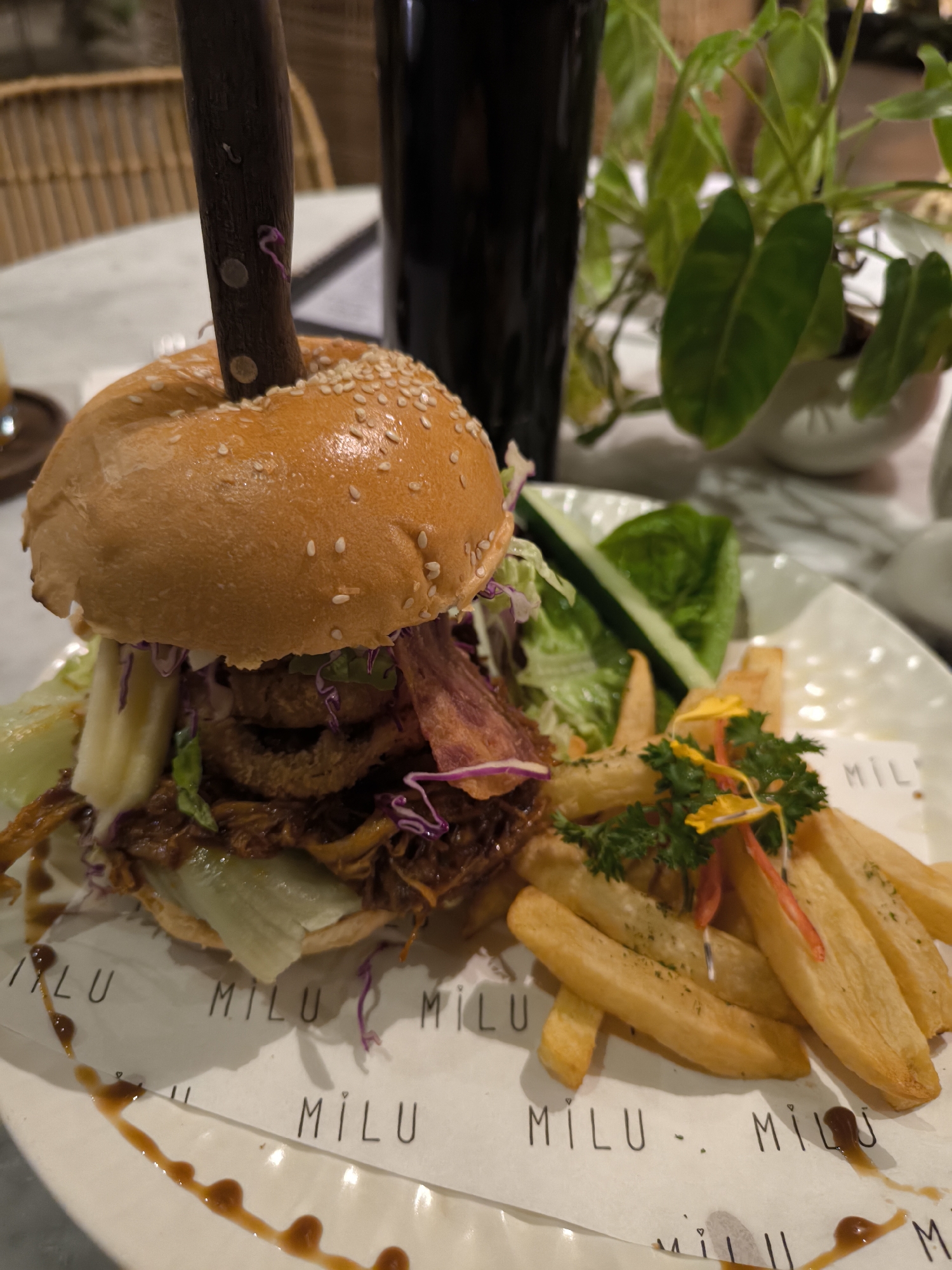 This close-up, eye-level shot captures a gourmet burger meal served on a white ceramic plate with a branded paper liner. The scene is set indoors, possibly in a restaurant or cafe in North Kuta, Indonesia, hinted at by the background elements.
The main subject is a generously sized burger, held together by a dark-handled steak knife impaled through its center. The burger features a sesame seed bun, pulled meat (likely pork or beef), crispy onion rings, shredded red cabbage, and fresh green lettuce. A creamy sauce is visible within the layers.
Accompanying the burger is a side of golden-brown french fries, garnished with fresh green parsley and small yellow and red edible flowers or peppers, adding a pop of color. A small, simple side salad with lettuce and a sliced cucumber strip is also present. A rich, dark sauce is artfully drizzled on the plate next to the burger and fries.
In the blurred background, a dark-colored glass or bottle is visible on the left, and a vibrant green potted plant sits on the right, suggesting a natural and relaxed ambiance. Part of a wicker-style chair is faintly discernible behind the plant, reinforcing a casual dining setting.
The lighting suggests an indoor environment, possibly evening, with warm tones illuminating the food. The paper liner beneath the food prominently displays the word "MILU" several times in a stylish font. The overall presentation is appealing and indicates a well-prepared meal.