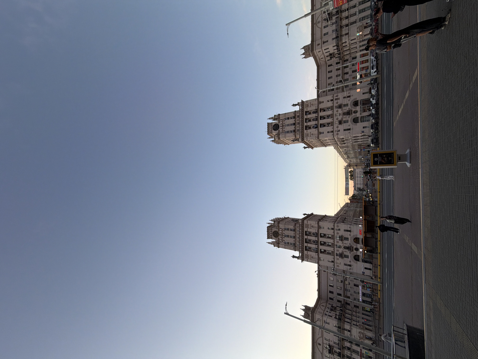 The image depicts a wide urban street scene in Minsk, Belarus, during the late afternoon or early evening. The perspective is slightly low-angle, showcasing a vast expanse of clear sky above grand, Stalinist Empire-style architecture.
Two imposing, light-colored buildings with multiple stories, detailed facades, and distinctive clock towers dominate the right side of the frame, flanking a broad avenue. These structures are rich in architectural details, including numerous windows and ornate cornices.
The street itself features a wide paved area with a sidewalk on the right and a road with visible lanes. Several individuals are present: a prominent pair, a man and a woman in dark clothing, are standing in the mid-ground on the sidewalk. Other pedestrians and vehicles, both parked and moving, can be seen further down the street, indicating a bustling urban environment. A streetlight pole is visible in the mid-ground.
The sky transitions from a light, slightly yellowish hue near the horizon, suggesting the approach of sunset or recent sunrise, to a clear, deep blue overhead. A few faint, wispy clouds are discernible in the upper left portion of the sky. The weather appears fair and calm.
A prominent "KFC" sign is visible on one of the buildings, indicating a commercial establishment. A street-side advertising stand is also present on the sidewalk, though its specific content is not legible. The scene conveys a sense of quiet grandeur in a historic city setting as daylight wanes.