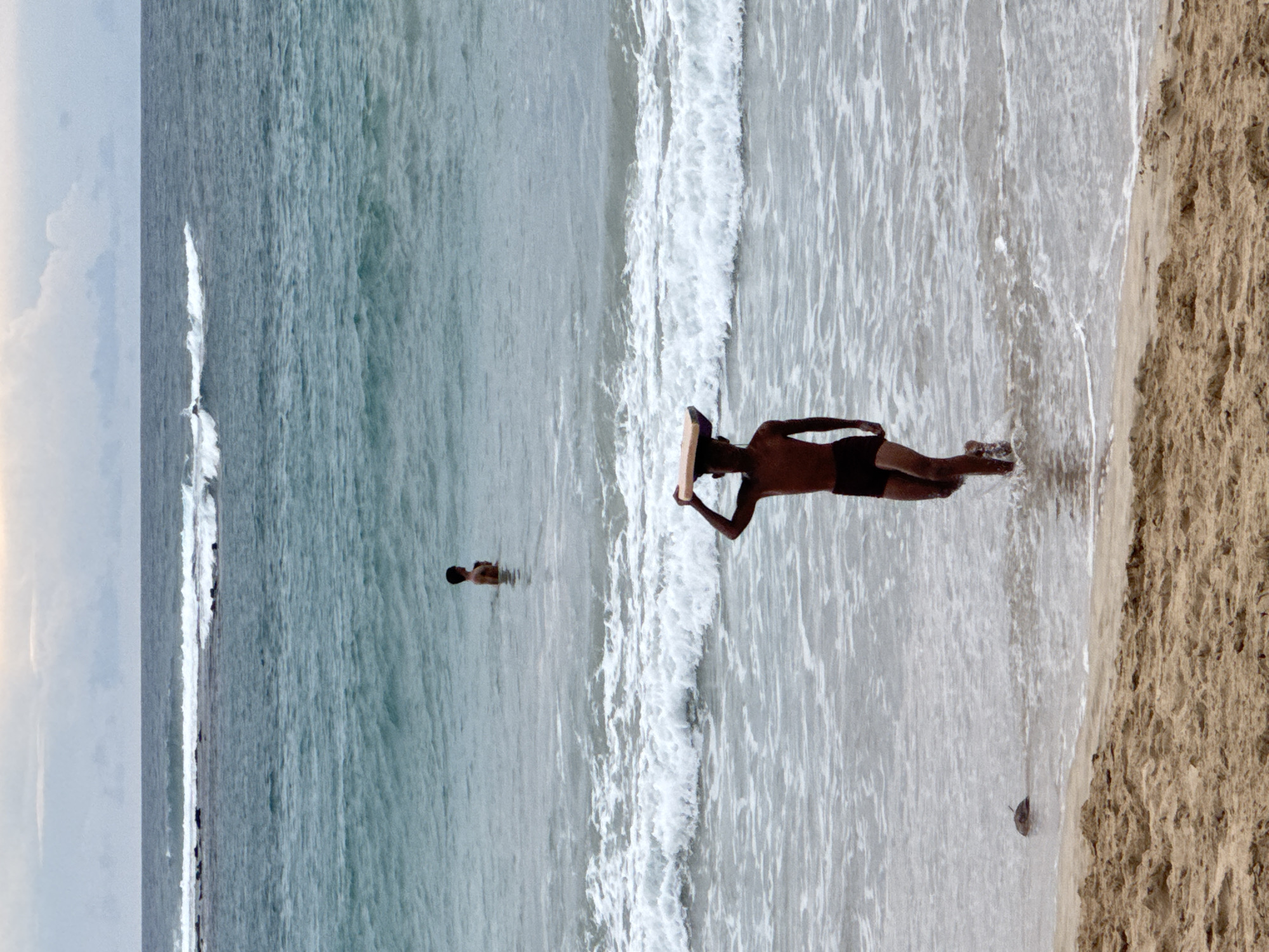 A young boy walks out of the ocean on a beach in Meunasahbale, Indonesia, carrying a light brown, flat object on his head. He is wearing dark swim shorts. His feet are submerged in shallow water, and ripples extend outwards from his legs. Behind him, another boy stands in the water, his body visible up to his chest. The ocean is a deep turquoise color with white waves crashing closer to the shore. The sky above is overcast with pale clouds. The foreground of the image is a sandy beach with visible footprints and small debris.