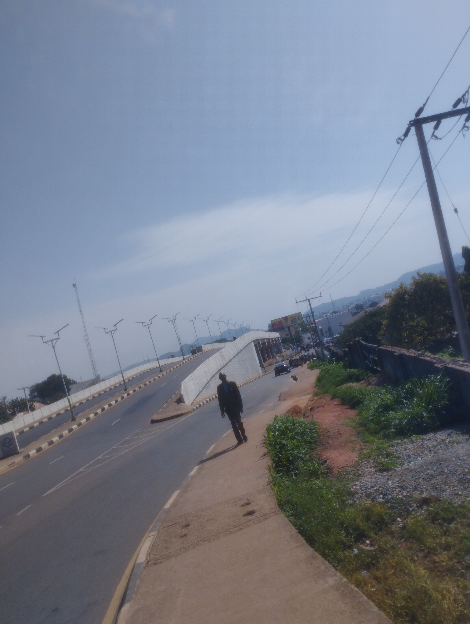 The image depicts a street scene in Jos, Nigeria, likely a highway overpass. The sky is clear and blue with some faint clouds. A man in a dark suit and pants is walking on the right side of the sidewalk, away from the camera. To his left is a multi-lane road with a divided highway, featuring streetlights and concrete barriers. The road appears to be an elevated section or overpass, with traffic visible in the distance. The surrounding environment includes some vegetation, a retaining wall, and buildings in the background. A utility pole with electrical lines is prominent on the right side of the frame. The overall impression is a typical urban landscape in Jos, characterized by modern infrastructure and daily activity.
