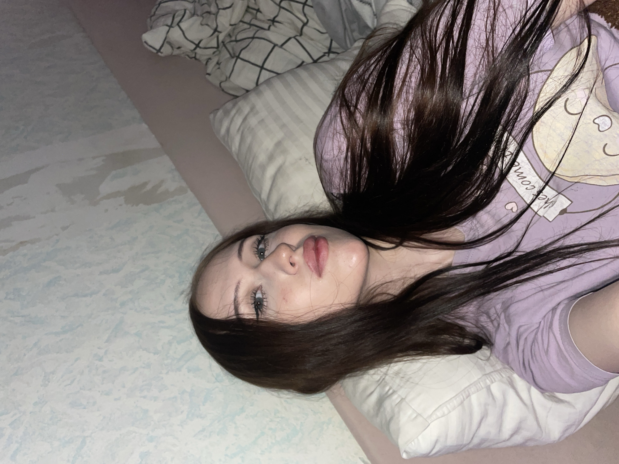 The media file shows a young woman lying on a bed, with her head resting on a pillow. She has long, dark hair and is wearing a purple t-shirt with a graphic print. The print features a crescent moon, stars, and the text "Welcome". The setting appears to be indoors, likely a bedroom, given the presence of a bed and pillows. The lighting suggests it is nighttime or a dimly lit room. The woman's facial expression is relaxed. The image is oriented vertically.