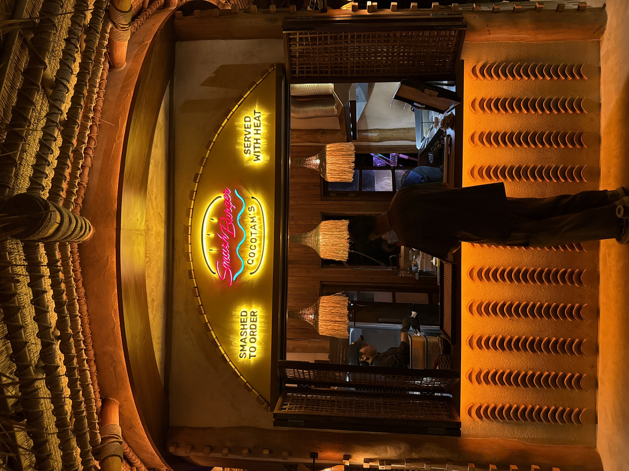 The image depicts the exterior of a food stall in Baan Bo Phut, Thailand, at night. The stall has a rustic, tropical-themed design, with a thatched roof and wooden accents. A prominent neon sign above the serving window reads "Smash Burger" in pink and "COCOTAM'S" in yellow, with the phrases "SMASHED TO ORDER" and "SERVED WITH HEAT" in yellow neon to the left and right respectively. The serving window is open, revealing a glimpse of the interior where at least one staff member is visible preparing food. Three decorative lamps with fringed shades hang from the ceiling above the serving area. In the foreground, a person stands with their back to the camera, facing the stall. The lower part of the stall features a textured wall illuminated by warm orange lighting. The overall atmosphere is cozy and inviting, suggesting a popular local eatery.