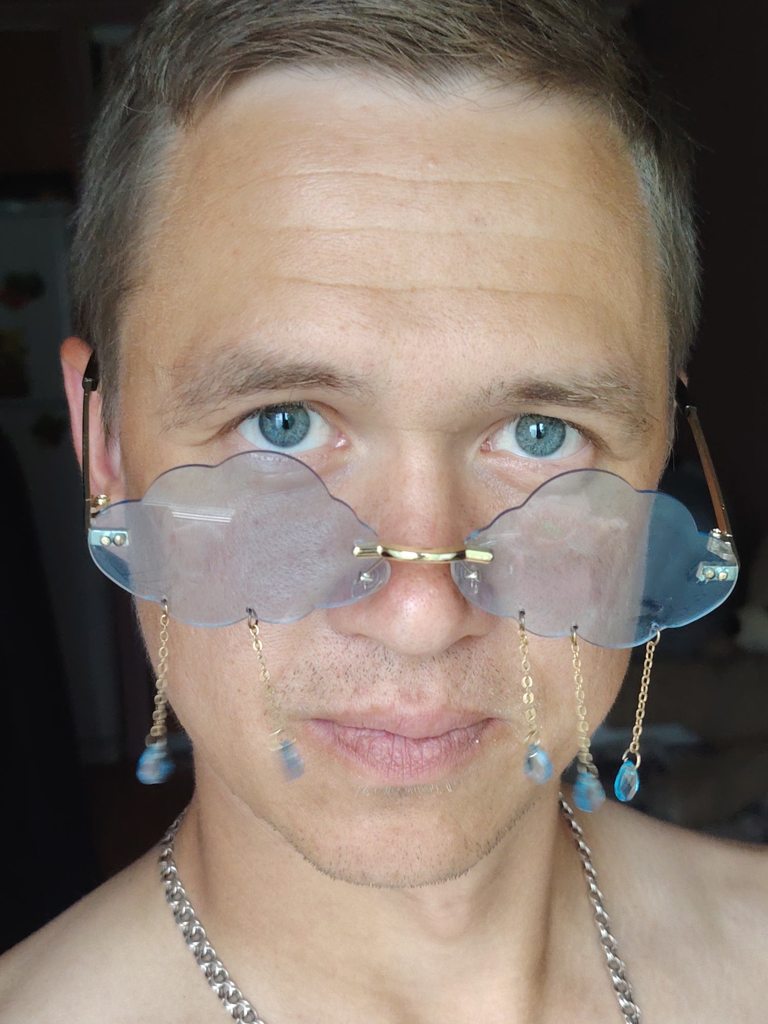 A close-up shot features a person's face, wearing novelty eyeglasses shaped like clouds. The glasses have gold accents and are adorned with delicate gold chains from which blue, teardrop-shaped beads hang. The person has blue eyes and is wearing a silver chain necklace. The background is dark and blurred, providing no discernible details about the environment. There is no information or visual cues to indicate the image is from Chaikovsky, Russia.