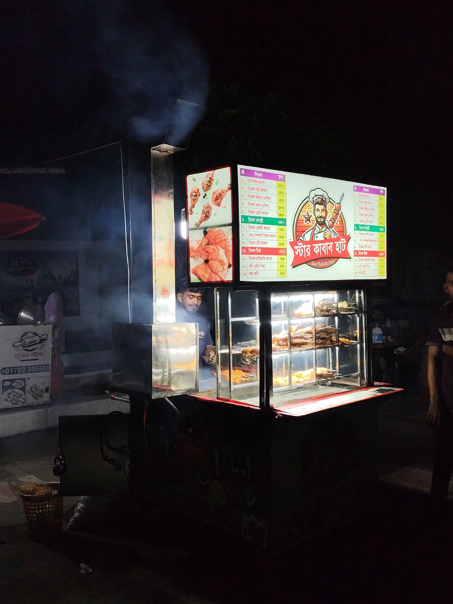 This is a nighttime, eye-level, medium shot of a food stall in Dhaka, Bangladesh. The stall is illuminated by overhead lights and a bright sign advertising "Star Kebab Hut." The sign displays images of chicken and a cartoon chef holding kebabs. A menu is also visible on the sign, written in Bengali script.
In the foreground, a glass display case showcases various types of kebabs on skewers, ready to be cooked. Behind the display case, a man with dark hair and a beard, wearing a dark blue t-shirt, is tending to the food. He appears to be a vendor or cook. Smoke is rising from a metal vent above the stall, suggesting that food is actively being cooked.
To the left of the food stall, there is a white cart with a rolling pin graphic and text that reads "01762 305500". A wicker basket filled with food items is placed near this cart. The overall environment appears to be an outdoor, possibly street-side, setting, indicated by the darkness and the presence of a sidewalk or road. Another person, whose face is not clearly visible, is partially seen on the right side of the frame, standing near the stall. The lighting creates strong contrasts and shadows, typical of a nighttime scene.