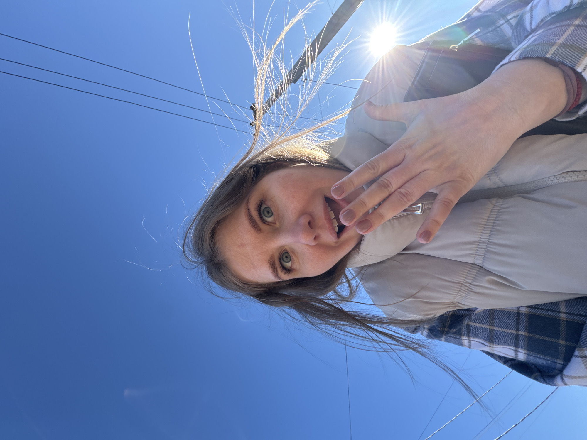 A woman in Горный, Russia, is captured in a low-angle selfie against a backdrop of a clear blue sky and power lines. The sun is visible, creating a bright glare. She has fair skin, green eyes, and light brown hair. She's wearing a light-colored puffer vest over a blue and white plaid shirt. Her hand is positioned near her face, with visible painted fingernails.
