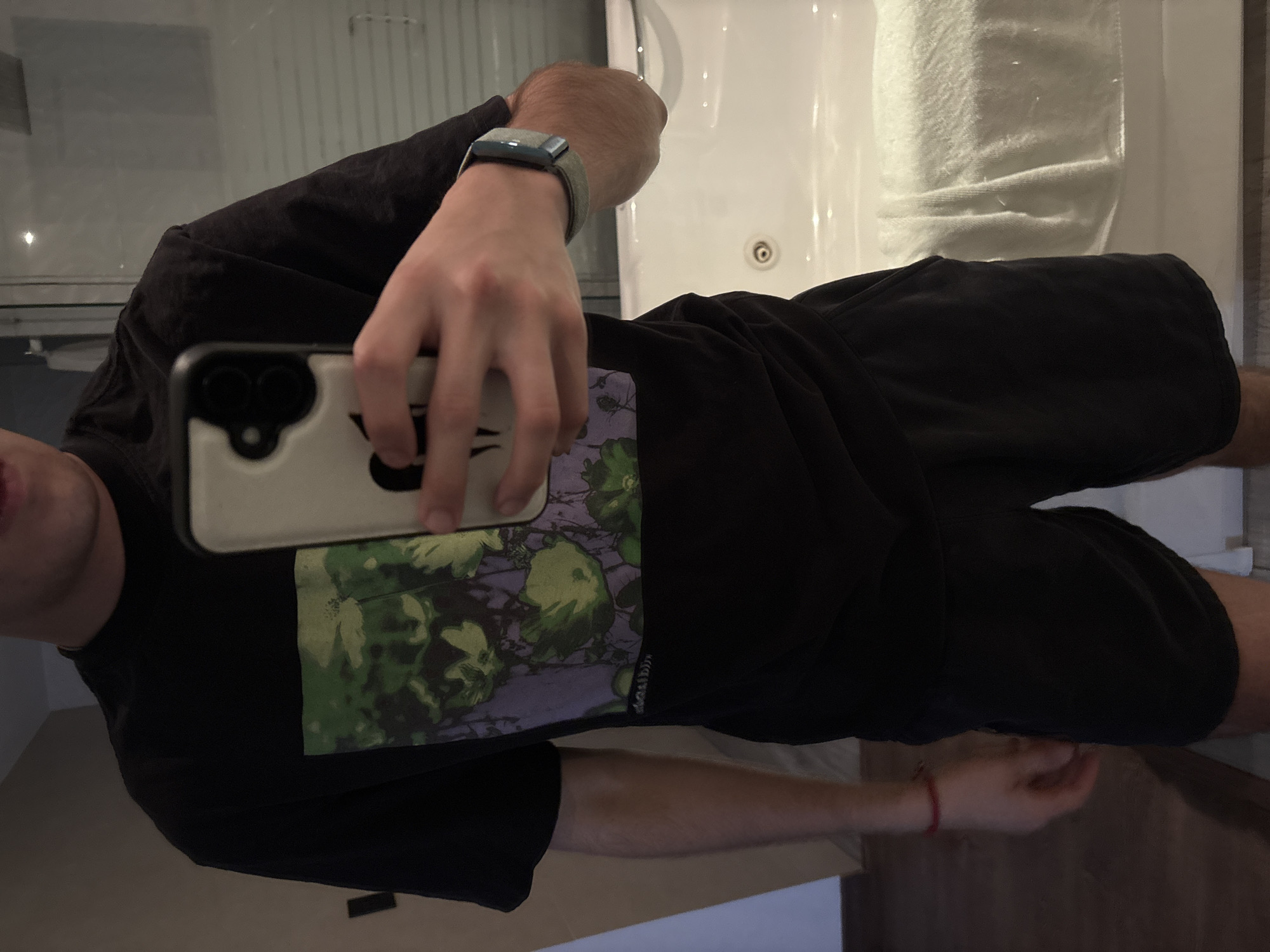 A person is taking a mirror selfie in what appears to be a bathroom. The person is wearing a black t-shirt with a green and purple graphic design, black shorts, and a smart watch with a green wristband. The phone being used for the selfie has a light-colored case. In the background, there's a bathtub and a partially visible towel. The location, San Bernardino Tlaxcalancingo, Mexico, cannot be verified from the given image.