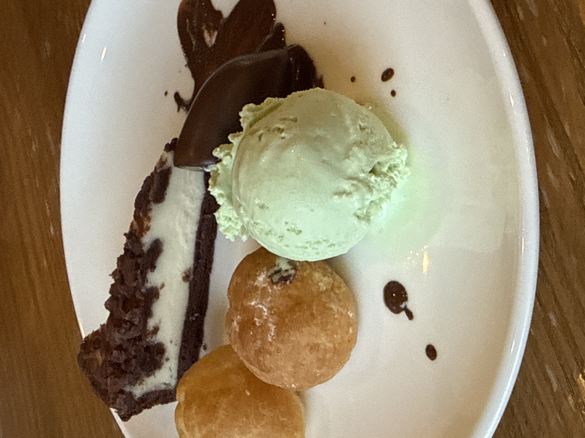 A close-up, overhead shot shows a white plate on a wooden table, featuring a dessert with several components. The dessert includes a slice of chocolate cheesecake with a dark crust and white filling, topped with crumbled chocolate. To the left of the cheesecake are two small, golden-brown fried dough balls. Adjacent to these is a scoop of pale green ice cream, likely pistachio or mint. A dark brown, curved chocolate shard or piece of fudge sits to the right of the ice cream, partially submerged in a smear of chocolate sauce. Small droplets of chocolate sauce are scattered across the plate. The lighting is natural, highlighting the textures of the food. The scene is indoors, likely a restaurant or cafe, with the wooden grain of the table visible in the foreground. The image does not provide any information about the city of Nordhausen, Germany.
