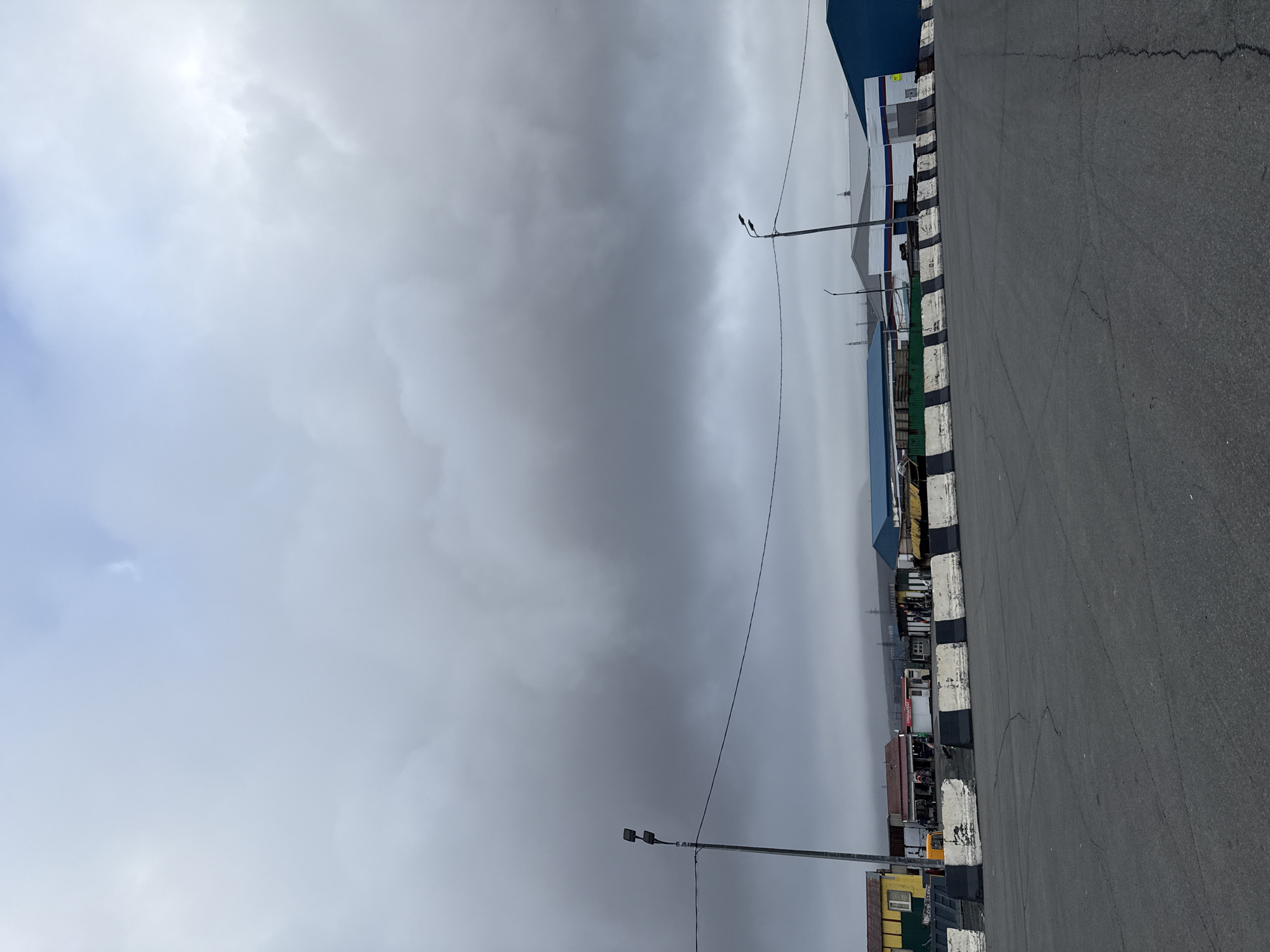 This is an overcast, wide-angle shot of an outdoor area in Norilsk, Russia. The sky is dominated by heavy, grey clouds. In the foreground, a dark asphalt surface shows cracks and is bordered by a low concrete barrier painted with alternating black and white stripes. Behind the barrier, a row of industrial or commercial buildings is visible. To the left, a small yellow building with a red roof sits next to a larger building with a red roof and what appears to be a sign. Further to the right, there are several industrial structures with metal siding, some painted in blue, white, and red stripes. A single lamppost with two lights stands tall on the left side of the image, with a wire stretching across the frame. Another lamppost is visible on the right. In the background, distant hills are obscured by fog or low clouds. There are no visible people in the image. The overall impression is one of an industrial, possibly somewhat desolate, urban or port setting under a grim sky.