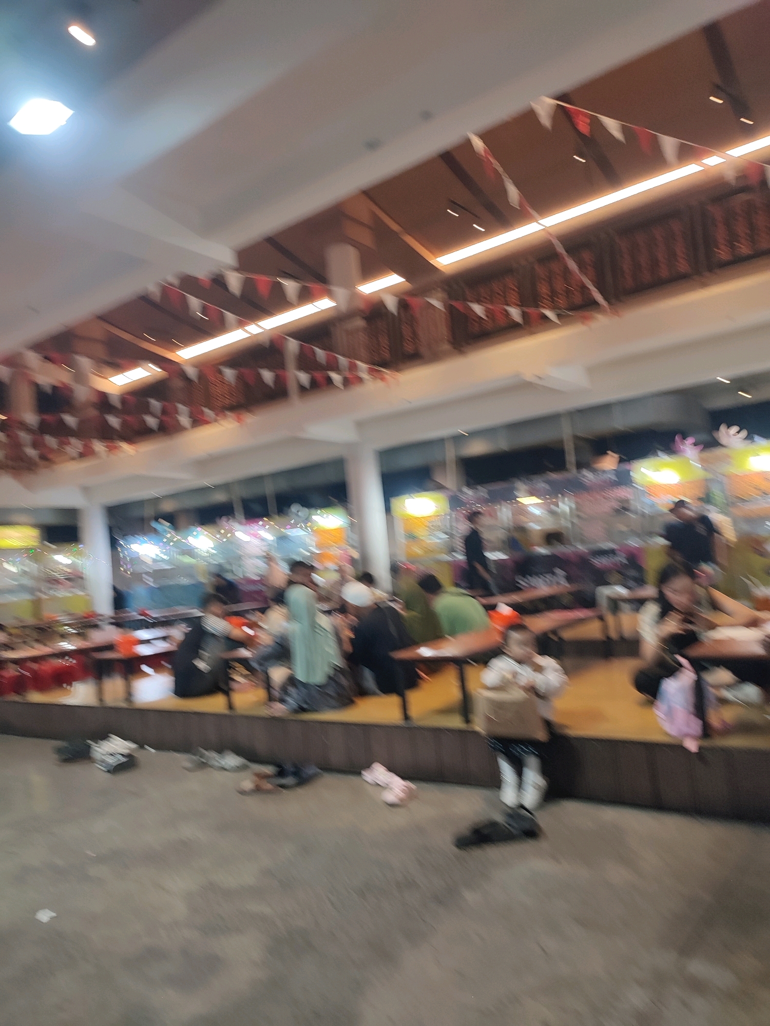 This image captures a bustling indoor marketplace or food court in Yogyakarta, Indonesia, possibly during a festive occasion given the red and white triangular flags decorating the ceiling. The scene is characterized by a warm, ambient lighting from overhead fixtures and illuminated stalls in the background, which are difficult to discern clearly due to motion blur and low resolution.
In the foreground, the concrete floor shows scattered footwear, suggesting a casual and relaxed atmosphere where people might remove their shoes. Several tables and benches are arranged on a raised wooden platform, occupied by patrons. People are seen sitting, eating, and socializing. A young child in a white top and patterned skirt is prominently featured, holding a bag. Other individuals in the mid-ground appear to be dressed in casual attire, with some wearing head coverings, indicating a mixed crowd.
The background reveals rows of stalls with colorful displays and lighting, hinting at a variety of vendors and products. The overall impression is one of a lively gathering place, likely a popular spot for locals to dine and shop. The specific location within Yogyakarta is not identifiable from the image, but the presence of the Indonesian flag colors and typical market architecture suggests it is a local establishment.