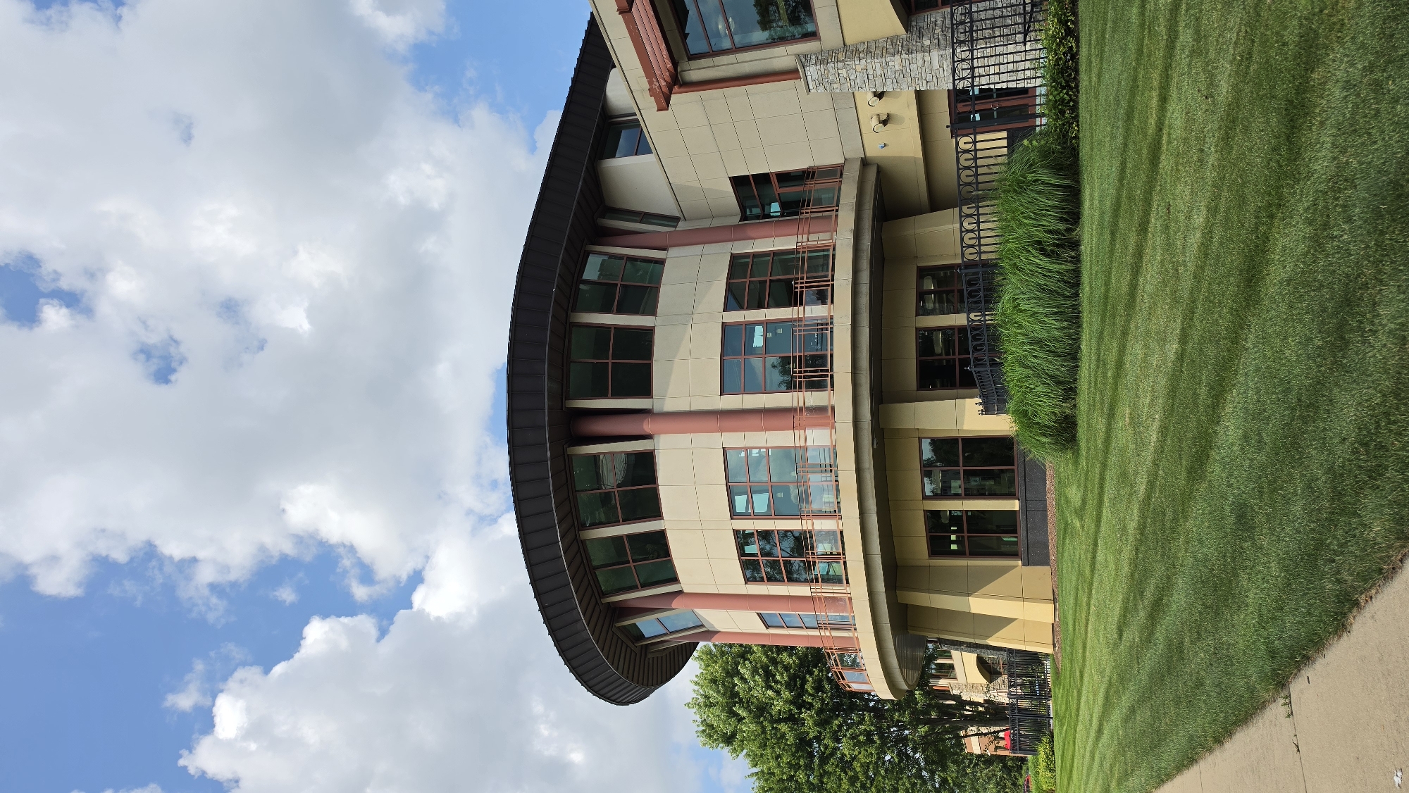 This is a medium shot, eye-level, exterior, daytime photograph of a modern building with a curved facade in Elgin, United States of America. The building is predominantly light beige with a dark brown, curved roof. Reddish-brown pillars support the upper level, which features large, multi-paned windows with dark frames. A balcony with a reddish-brown railing wraps around this upper section. The lower level of the building also has windows and is a lighter beige color, with some sections appearing to be yellow. A stone-clad column is visible on the right side of the building. In the foreground, a neatly mowed green lawn with visible mowing stripes stretches across the frame. Behind the lawn, a decorative black metal fence encloses a patch of tall, ornamental grass. A concrete sidewalk runs along the left edge of the image. Lush green trees are visible to the left of the building. The sky above is blue with scattered white, fluffy clouds.