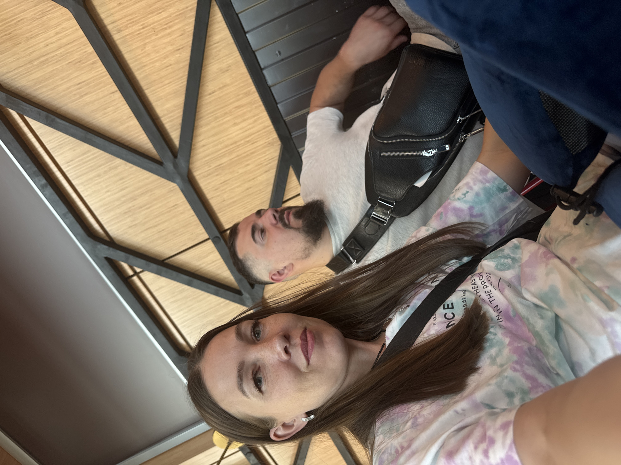 The image captures two individuals, seemingly in a casual setting. A woman with long brown hair is taking a selfie, with a man lying behind her. The man has a beard and is wearing a white t-shirt and a black crossbody bag. They are positioned near a structure with a geometric pattern made of wood panels and dark metal framing. Part of a blue object is also visible in the background. The clothing and accessories suggest a contemporary style. Based on the text on the woman's shirt, they are located in Bol'shie Kabany, Russia, specifically at coordinates E 49.62 km.