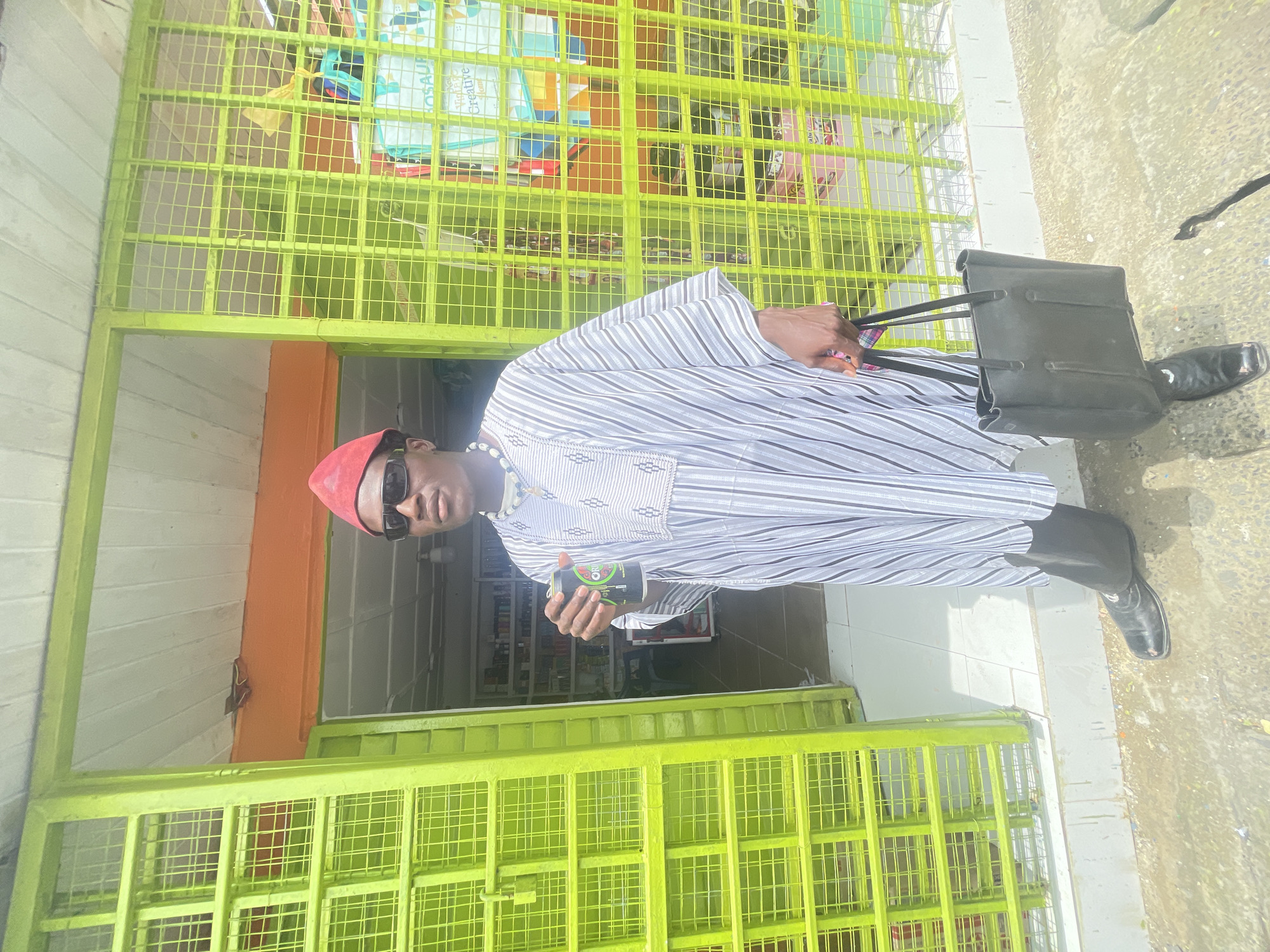 A man in Ado, Nigeria stands in front of a store. He is wearing a red cap, sunglasses, and a striped robe. He is holding a black bag and a drink. The store has a green barred window.