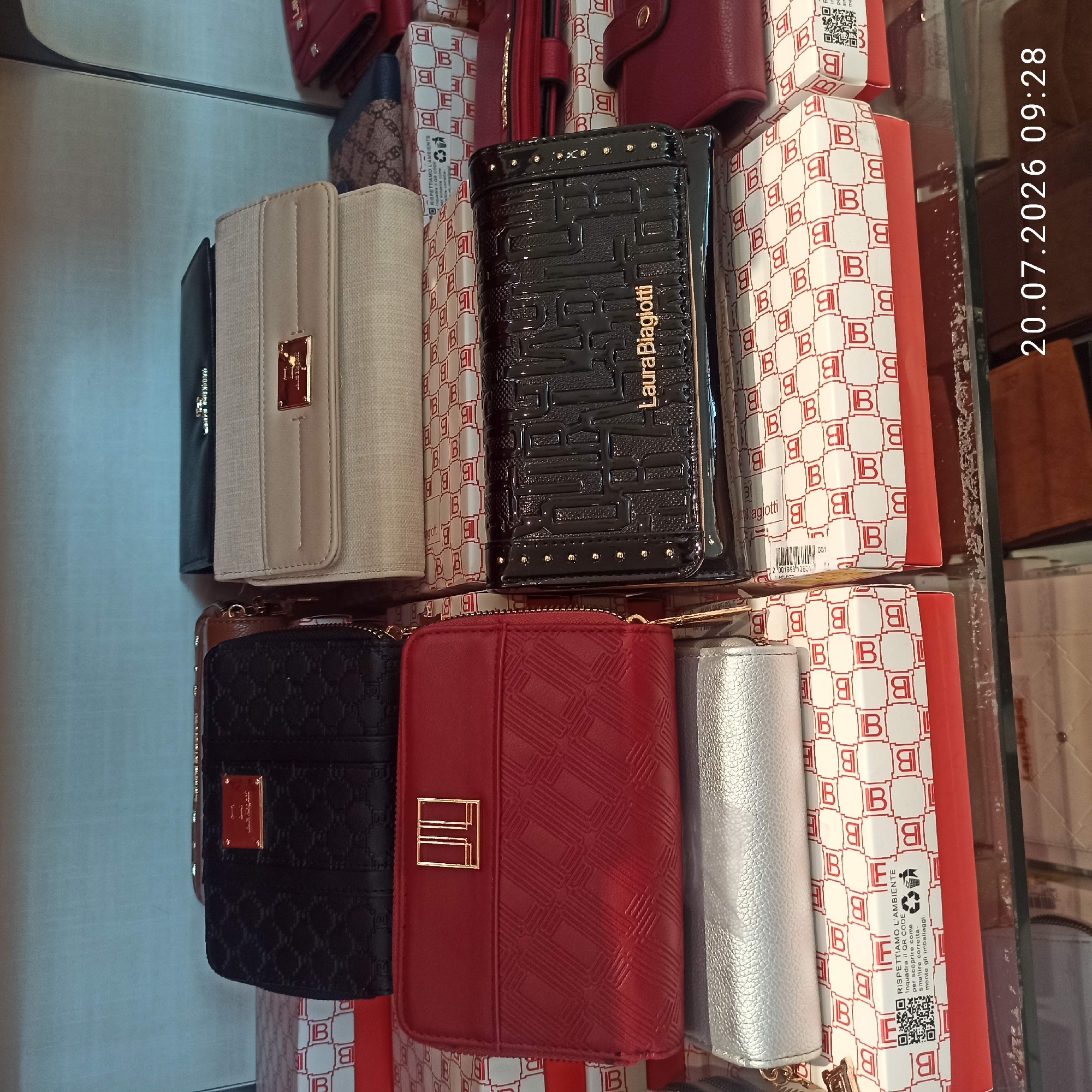 The image displays a retail store shelf filled with women's wallets and clutch bags. There are multiple items stacked on top of each other and arranged side by side. The wallets are in various colors including black, red, beige, and silver. Some of the wallets feature embossed patterns and metallic brand logos. The boxes the items are in are also visible, some with a repeating "B" logo pattern and some with recycling symbols and QR codes. A date stamp "20.07.2026 09:28" is present in the bottom right corner of the image. There are no people visible in the image. The background appears to be a light blue wall. The items are organized in a retail display setting.