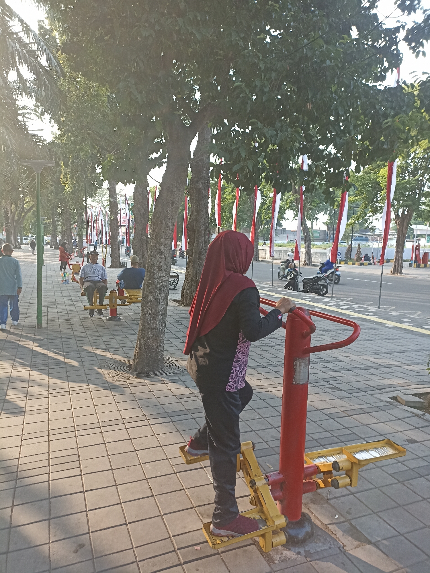 In Kelurahan Kepatihan, Indonesia, on a sunlit day, a woman wearing a maroon hijab and dark athletic clothing is using a red and yellow outdoor fitness machine, specifically an elliptical trainer, with her right foot placed on the pedal. She is standing on a paved walkway made of interlocking square tiles, casting long shadows from the trees lining the street. Behind her, the street is visible with several motorcycles passing by, and red and white Indonesian flags are hung along the roadside. Other people are scattered throughout the scene, some sitting on similar yellow exercise equipment, and others walking along the paved path. The environment is urban, with trees providing shade and a clear sky suggesting fair weather.