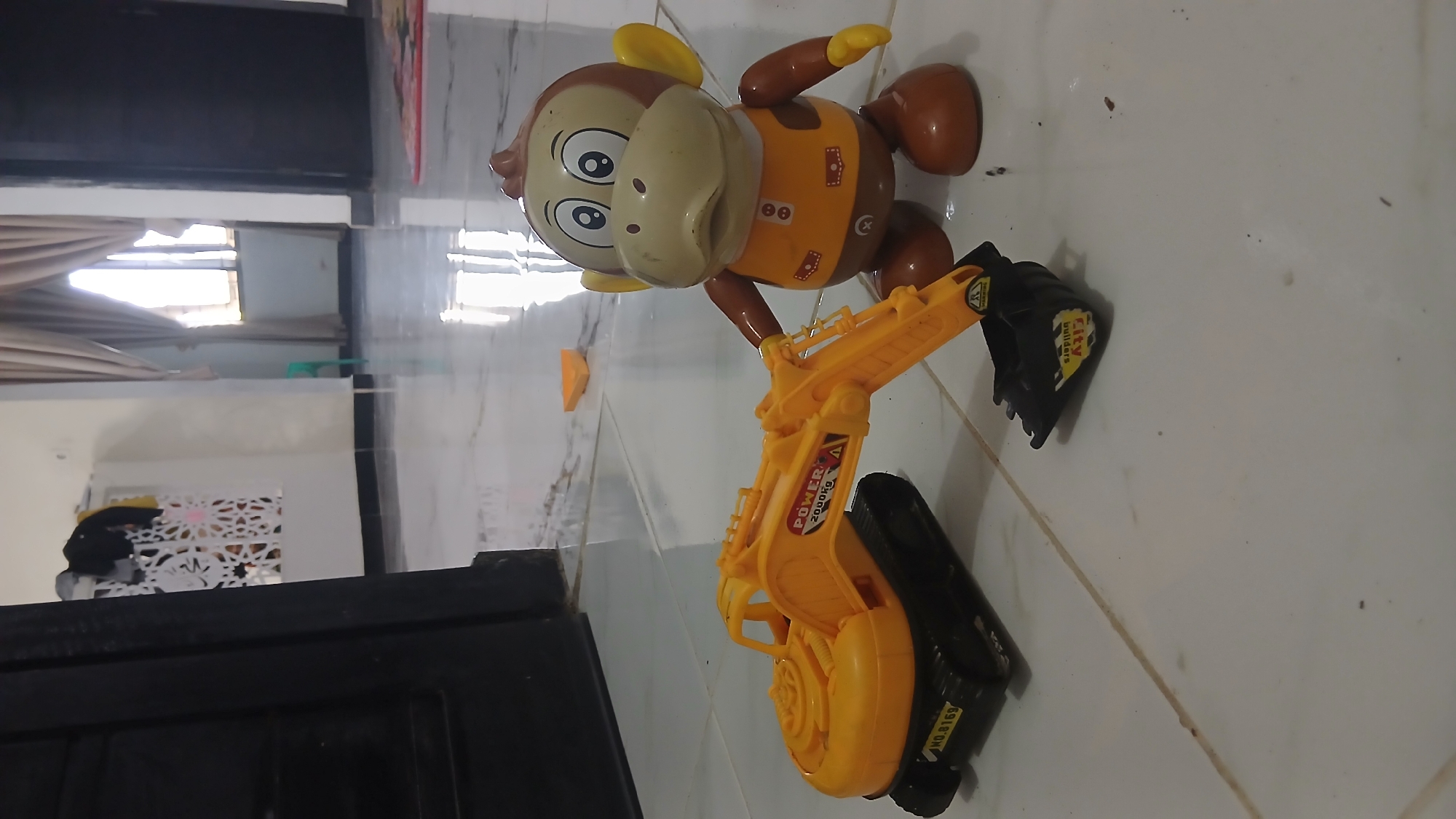 The image shows a toy excavator and a toy monkey on a tiled floor. The excavator is yellow with black tracks and a black bucket. The monkey is brown and beige with large eyes and ears, wearing an orange vest. The toys are arranged as if the monkey is operating the excavator. The background is a room with white walls, a window with beige curtains, and a dark doorway. A patterned screen is visible on the left side of the frame. The lighting suggests it is daytime, with sunlight coming through the window. The scene is indoors, likely in a residential setting.
