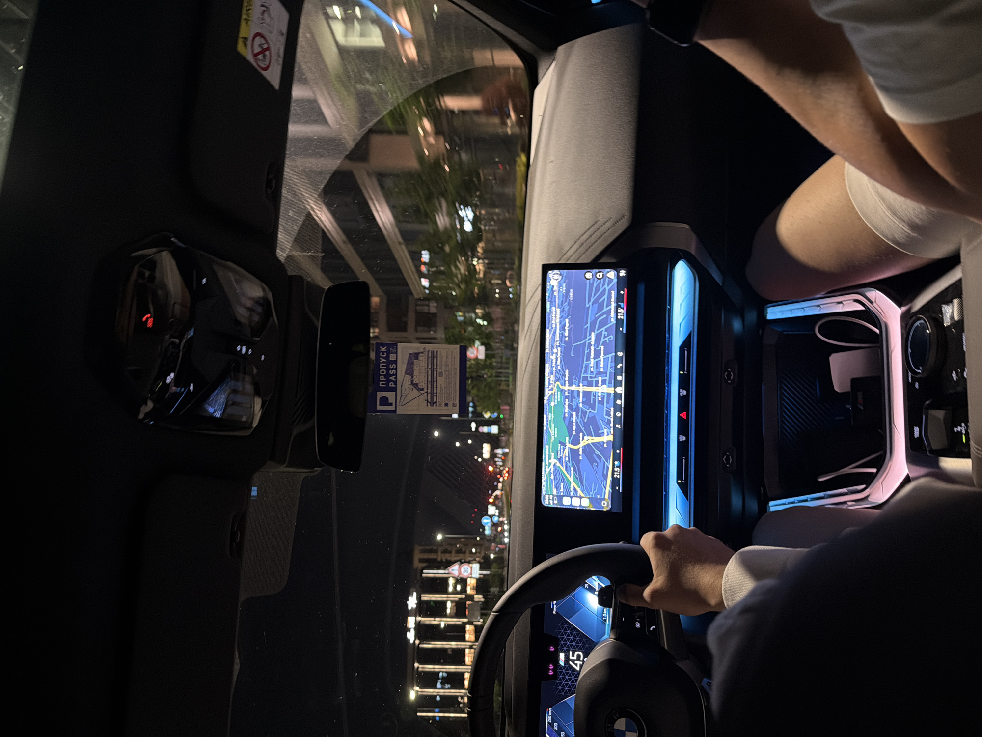 The image captures the interior of a modern BMW vehicle, likely an electric model, at night, looking forward through the windshield onto a city street in Sofia, Bulgaria.
In the foreground, a driver's right hand, wearing a light-colored long-sleeve top, grasps the steering wheel. The BMW logo is visible on the center of the steering wheel. To the left, a digital instrument cluster displays "45" (likely km/h), a speedometer indicating up to "260 km/h", and "1.0 km" for distance. A blue and white geometric pattern characterizes the display. To the right, a large infotainment screen displays a navigation map with a route highlighted in blue. Street names in Cyrillic, including "ул. Акад. Борис Стефанов" and "бул. Д-р Г. М. Димитров", are visible on the map, confirming the location as Sofia, Bulgaria. The screen also shows "21.5°C" and music/media icons.
Blue ambient lighting illuminates the dashboard and the area beneath the main screen. Further down, a wireless charging pad area is illuminated with pink/purple light, containing a white cable and displaying an "M" logo. A passenger's right leg, clad in shorts, and a forearm are visible in the lower right corner of the frame.
Above the dashboard, a rearview mirror is present. Hanging from it is a blue and white paper pass with the Cyrillic text "ПРОПУСК PASS" (meaning "Pass"). An "AIRBAG" warning label is affixed to the passenger-side sun visor. Overhead, a black roof liner and a central light/sensor console are visible.
Through the windshield, a multi-lane city street at night is visible. Buildings with illuminated windows line both sides of the road. Streetlights cast light onto the scene. In the distance, a circular red-bordered road sign indicating a "30" km/h speed limit is discernable. The overall impression is a vehicle in motion through a well-lit urban environment.