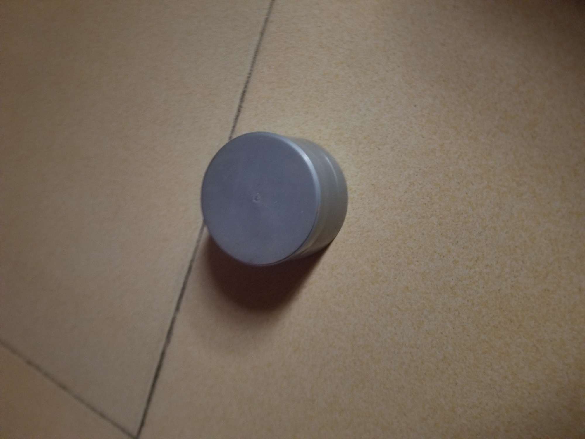 The image shows a close-up, overhead view of a small, gray, cylindrical object resting on a tiled surface. The tiles are a light brown or tan color and have visible grout lines that create a grid pattern. One prominent grout line runs diagonally from the bottom left to the top right of the frame, appearing to divide two larger tile sections. The cylindrical object is positioned slightly off-center to the left, casting a soft shadow to its right. The object itself has a smooth, matte finish and appears to be a cap or lid for something. There is a small circular indentation or marking on its top surface. The lighting suggests an indoor setting with overhead illumination, as indicated by the shadow. No people are visible in the image, and the scene is a simple close-up of an object on a floor. There is no visible text in the image.