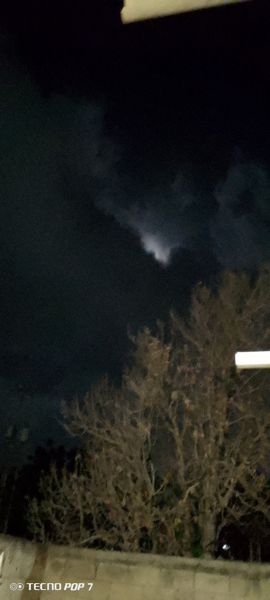 The image depicts a night scene in Altagracia, Venezuela. The sky is dark, with clouds partially obscuring what appears to be moonlight. In the foreground, there are bare tree branches and foliage. The bottom of the image shows a light-colored wall. A streetlight is visible on the right side of the image. The image was taken with a Tecno Pop 7 phone. No people are visible in the image.