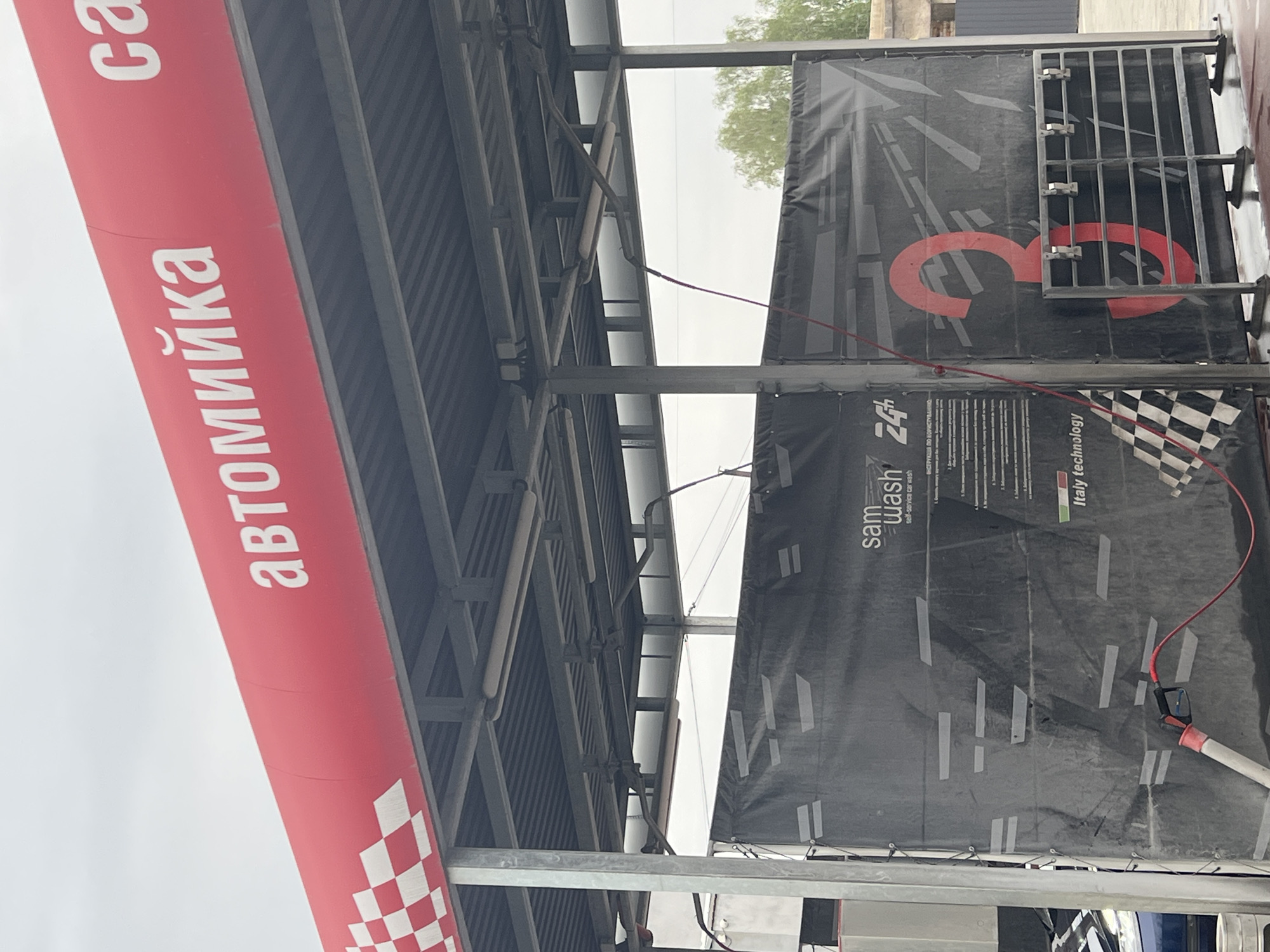 Here is a description of the image:
The image shows a car wash station in Horodok, Ukraine. The station has a red and white awning with the word "автомийка" written on it. There is also a banner with the words "sam wash 24" and "self-service car wash" written on it. The banner also has the words "Italy technology" on it. The image appears to be taken from under the awning of the car wash station.