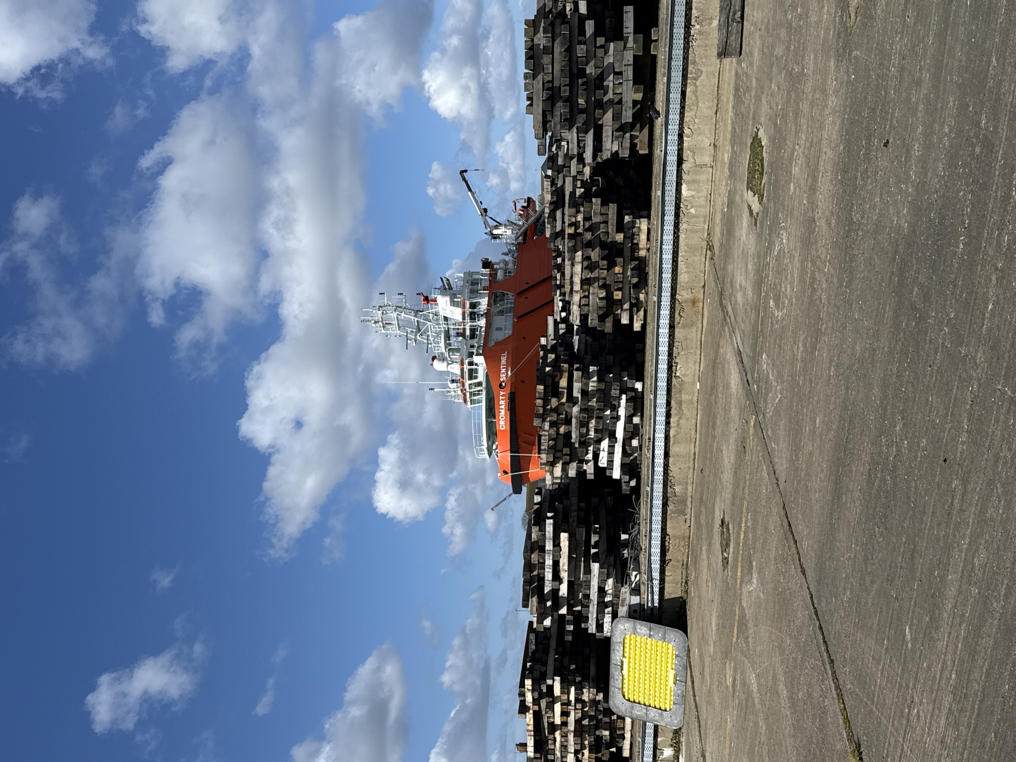 The image captures a static scene within the port area of Gorleston-on-Sea, United Kingdom, under a clear blue sky dotted with scattered white cumulus clouds. The foreground consists of a grey concrete ground surface, marked by linear cracks and small patches of green vegetation. A bright yellow, rectangular, ribbed safety mat or tactile plate with a dark grey perimeter is positioned on the concrete in the lower left quadrant. In the midground, several large, irregularly stacked piles of dark, weathered timber, consistent with railway sleepers, are arranged horizontally. These stacks partially obscure the background. Behind the timber, a concrete retaining wall supports a light blue/grey perforated metal conduit. Dominating the background is a large, orange-hulled vessel with a multi-level white superstructure, identified by the name "CROMARTY SENTINEL" emblazoned on its side. The vessel features multiple white antennae, masts, and a white crane boom extending from its deck. No individuals or explicit actions are visible within the frame.
