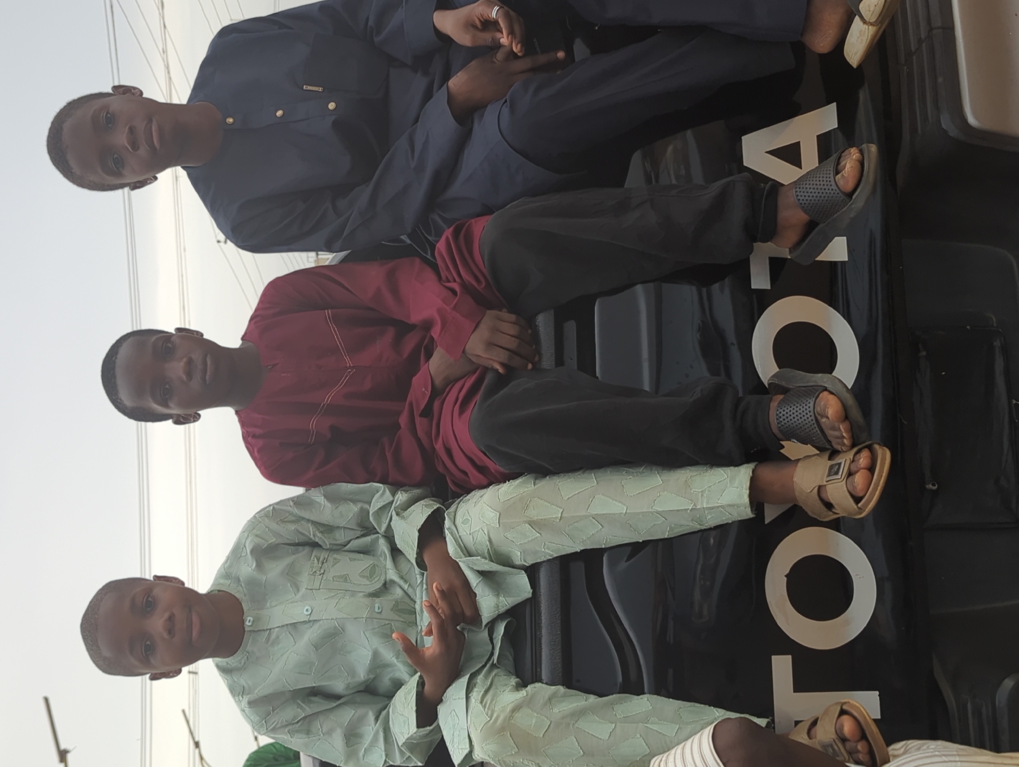 Three young men are sitting on the tailgate of a black Toyota pickup truck. The tailgate is marked with "TOYOTA" in large white letters. The truck is parked in an outdoor environment, likely in Kafanchan A, Nigeria. The men are sitting in a row from the front of the truck, and the camera is at a slight angle. The man in the front is wearing a light-green patterned robe and matching pants. The man in the middle is wearing a maroon-colored robe and black pants. The man in the back is wearing a dark blue robe and matching pants. All three men are wearing sandals.