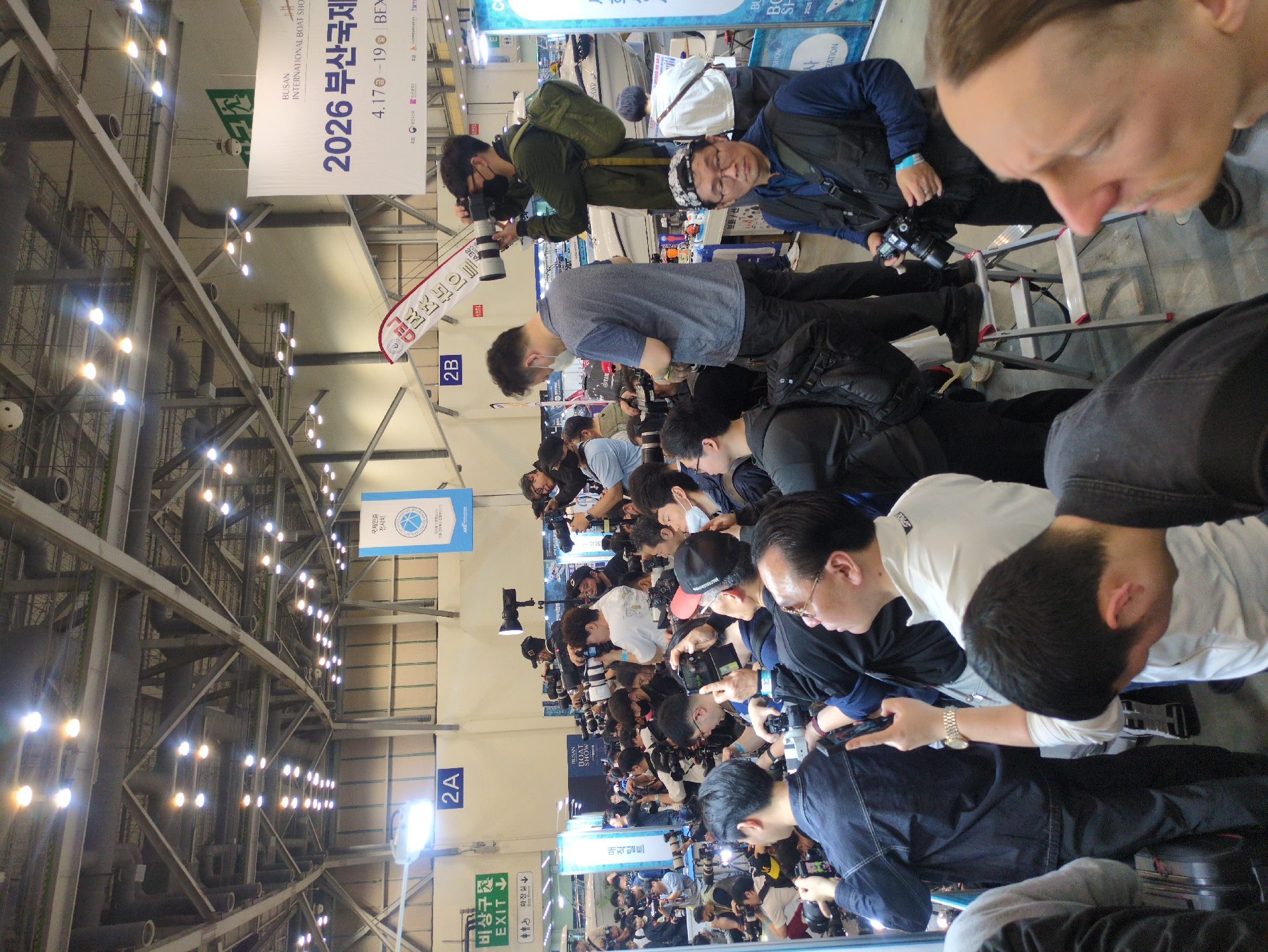 This image depicts a crowded indoor event, likely a press conference or exhibition, at a convention center in Busan, South Korea. The scene is dominated by numerous people, primarily men, clustered together, many holding professional cameras with long lenses or smartphones, actively taking photographs or observing something out of frame. A few individuals wear face masks. One man is positioned on a step ladder, also equipped with a camera, suggesting an effort to get a better vantage point.
The setting features a high industrial-style ceiling with visible metal trusses, air ducts, and rows of bright artificial lights illuminating the hall. In the background, sections of what appears to be a white boat or yacht are visible, aligning with the event's theme.
Prominent banners hanging from the ceiling provide key details about the event. A large white banner clearly reads "BUSAN INTERNATIONAL BOAT SHOW" and "2026 부산국제" (2026 Busan International), indicating the year, location (Busan, South Korea), and nature of the show. Dates "4.17 – 19" and "BEXCO" (a common exhibition center in Busan) are also visible. Other signs include "국제인증 전시회" (International Certified Exhibition), "비상구 EXIT," "화장실↓" (Restroom↓), and section markers "2A" and "2B." The overall impression is one of a busy, professionally covered event.
