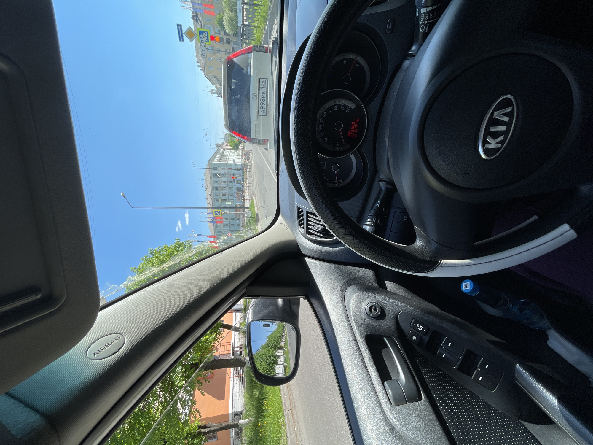 The image captures a view from the interior of a car, looking out through the windshield on a bright, sunny day. The car is stationary, with a white SUV directly in front.
**Subjects:**
Inside the car, a black steering wheel with a silver "KIA" logo is prominent on the right. The dashboard shows the speedometer at zero and an odometer reading "6069" km, with a trip meter at "1572.6" km. An "AIRBAG" label is visible on the A-pillar on the left. The driver's side door panel is also visible, featuring window controls and a bottle with a blue cap tucked into a compartment. The side mirror on the left reflects trees and a road, suggesting a sunny, tree-lined street.
Outside the car, the white SUV in front bears a Russian license plate "A998PK 124". Further down the street, buildings with windows line the road. Various road signs are visible, including a pedestrian crossing sign, a yield sign, and a "СТОП" (STOP) sign. A traffic light is also present. The sky is clear and blue, indicating fair weather.
**Activity or Event:**
The car appears to be stopped at an intersection or in traffic, waiting.
**Notable Details:**
The scene depicts daytime with bright sunshine. The Russian license plate and the "СТОП" sign confirm the location is in Russia. The provided context specifies the location as Zheleznogorsk, Russia.
**Visible Text:**
"KIA" on the steering wheel, "AIRBAG" on the A-pillar, "A998PK 124" on the license plate, "СТОП" on a road sign, and "TRIP", "MIST", "FRONT OFF" on the car's controls.