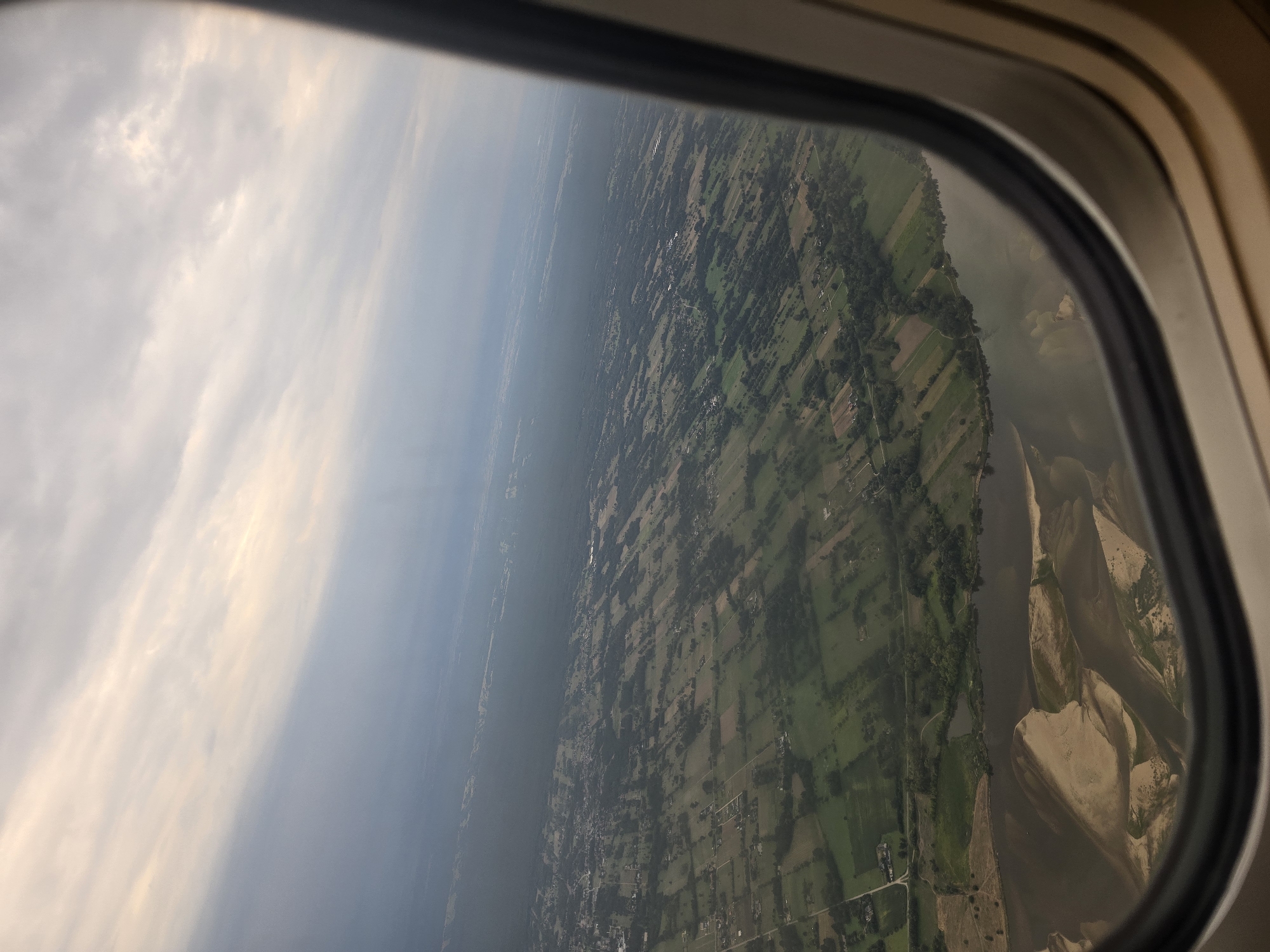 An aerial view, captured through an airplane window, shows a landscape composed of agricultural fields, a river, and a distant town. The sky above is overcast with clouds, tinged with the pale hues of either sunrise or sunset. The cultivated land is divided into rectangular plots, displaying varying shades of green and brown, suggesting different crops or stages of growth. A wide, dark river dominates the foreground, its banks showing patches of sand and vegetation. The town, visible in the middle ground, appears to be spread out with low-rise buildings and tree-lined streets. The overall scene is one of rural tranquility and vastness, a typical vista seen from an airplane during flight. There is no information available to identify this specific location as Kyustendil, Bulgaria.