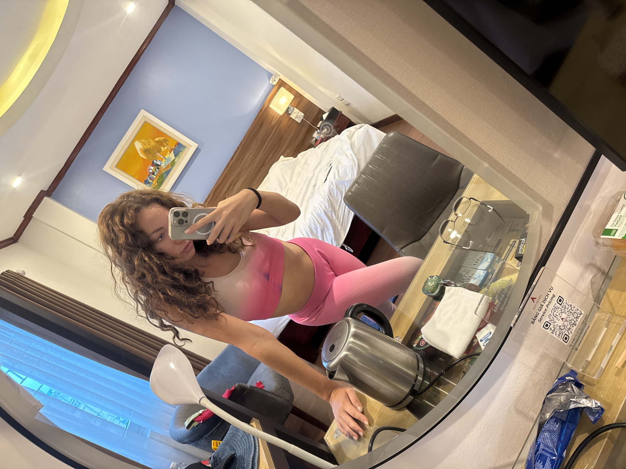 A woman with long, curly brown hair is taking a mirror selfie in a hotel room in Hanoi, Vietnam. She is wearing a pink sports bra and pink leggings. She is standing in front of a mirror, which reflects a hotel room with a bed, a desk with a kettle, and a painting on the wall. The room is lit by overhead lights and a bedside lamp.