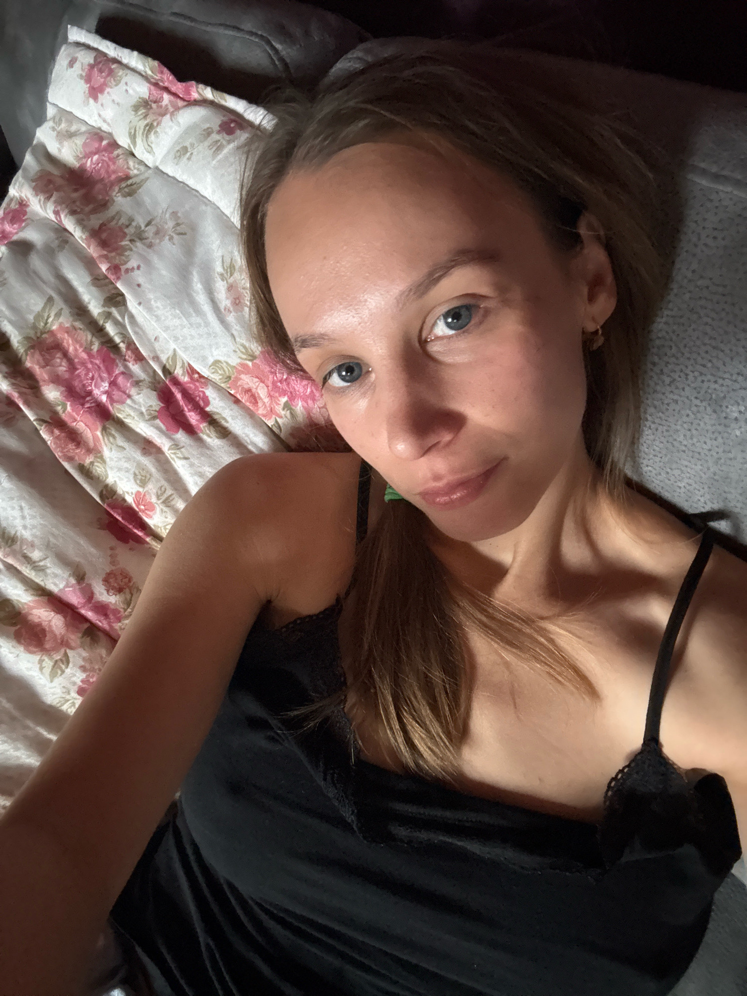 A close-up selfie shows a woman lying on her back, looking towards the camera. She has fair skin, blue eyes, and light brown hair that is slightly mussed. She is wearing a black camisole with lace trim. She is lying on a bed covered with a comforter that features a floral pattern with pink and white roses. The background shows a textured gray surface, possibly a couch or another blanket. The lighting is dim, casting shadows on her face and neck. The location is not specified in the image itself or the provided crops, therefore any mention of Überherrn, Germany cannot be substantiated.