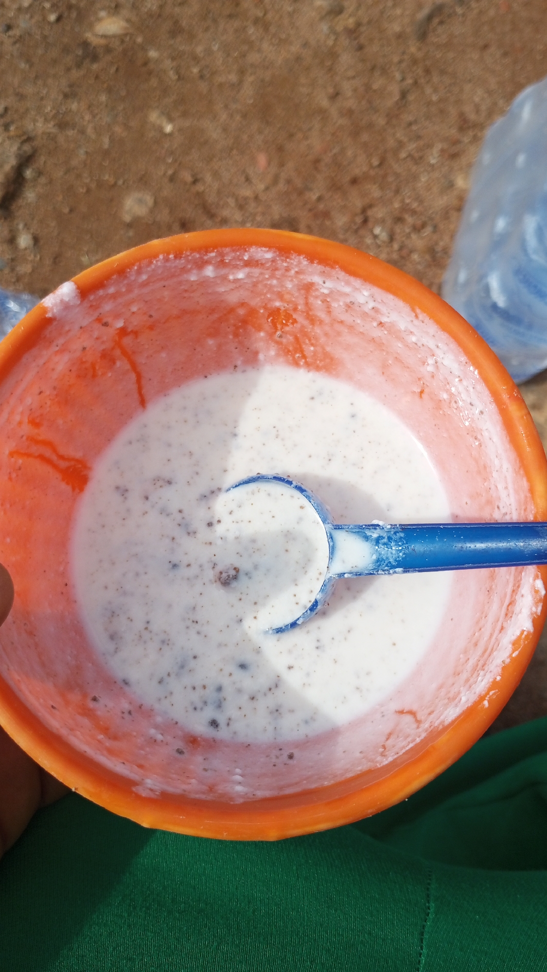 The image shows a close-up of an orange bowl filled with a white, creamy substance containing dark specks. A blue spoon is partially submerged in the substance. The bowl is on a surface that appears to be dirt or sand. In the background, to the right, a portion of a clear plastic bottle with blue contents is visible. The lower part of the image shows a green fabric. The overall impression is of a simple, perhaps rustic, setting, possibly outdoors. There is no specific visual information within the image that can confirm the location in Gura Top, Nigeria.