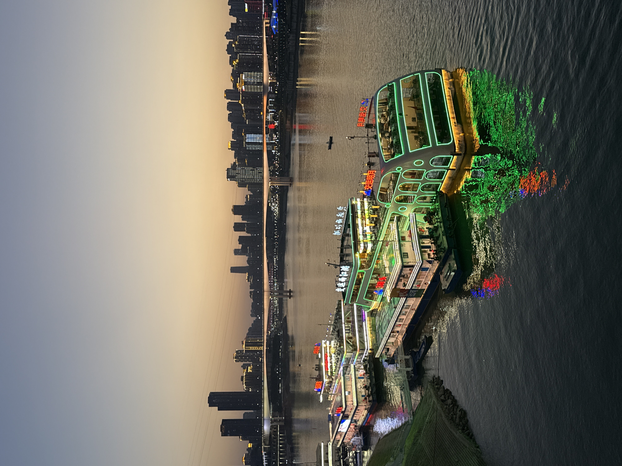 This is a twilight shot of multiple brightly lit tour boats docked on a wide river, likely the Yangtze River, in Yuzhong District, Chongqing, China. The boats are multi-decked and adorned with vibrant green and white neon lights that illuminate their structures and cast reflections on the dark water. Some of the boats have red Chinese characters displayed on their upper decks. In the background, a sprawling cityscape of tall buildings is visible, with many windows lit up. A large bridge spans the river, connecting different parts of the city. The sky is a gradient of pale blue and soft orange, typical of dusk. The water is calm, with gentle ripples on the surface. The overall scene depicts a lively urban waterfront at the transition from day to night, with the illuminated boats and city lights creating a dynamic and visually striking panorama.