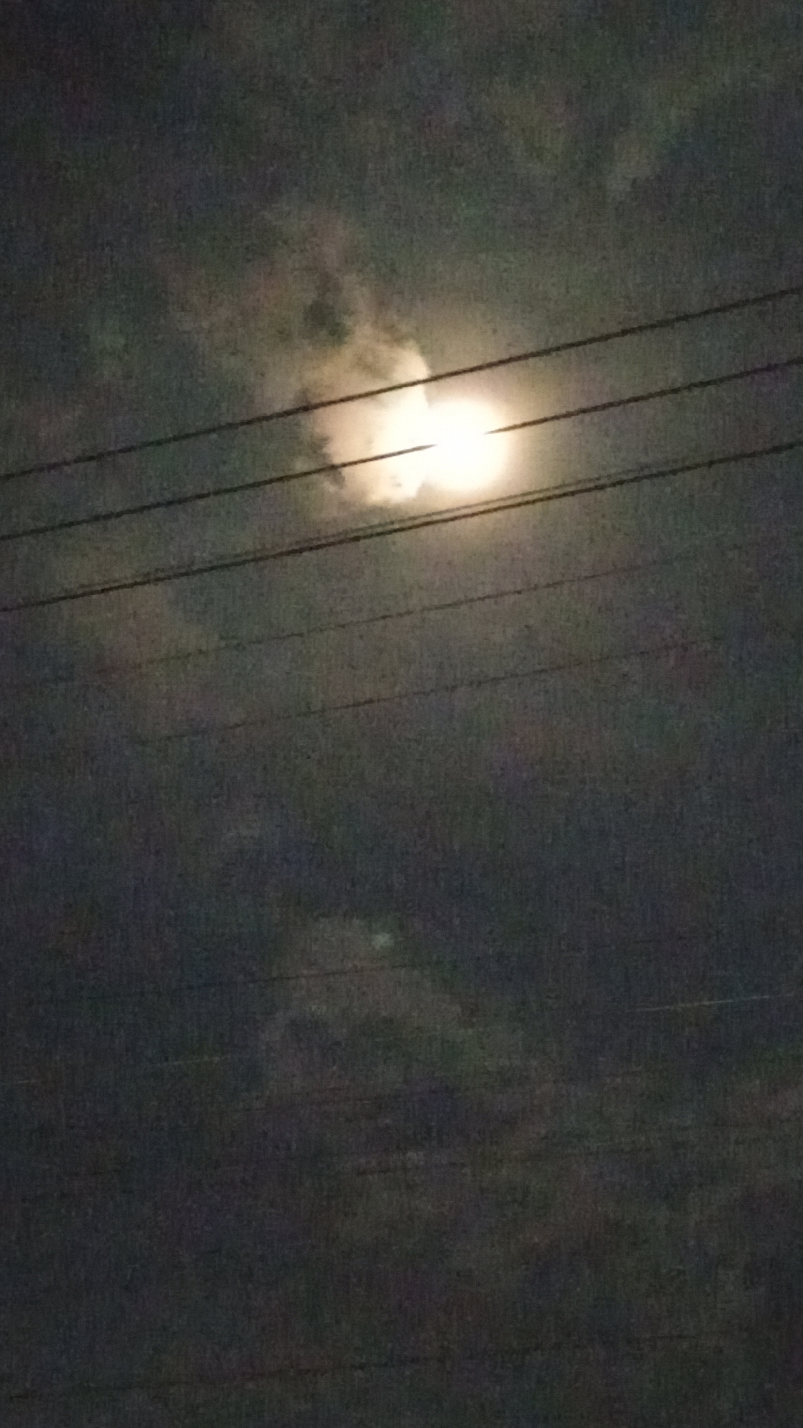 The image captures a nighttime sky with a bright, slightly obscured moon. Several horizontal power lines stretch across the frame, partially covering the moon and the surrounding clouds. The sky is predominantly dark, with patches of lighter clouds illuminated by the moonlight. The overall impression is of a cloudy night with a visible moon and utility infrastructure. No people or specific locations in Bireuenmeunasahcapa, Indonesia are discernible from the image.