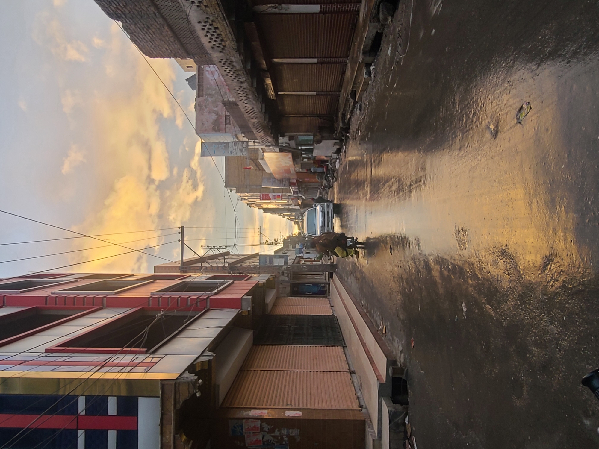 An eye-level, angled, long shot shows a wet road in Quetta City Tehsil, Pakistan, under a yellow and white sunset sky. On the left is a four-story building with a metal facade, painted red and white with blue accents. Across the road are old brick buildings with closed, corrugated metal doors. Overhead are electrical wires. In the distance, a white car moves along the road, and a person with a cart or wheelbarrow is also on the road.
