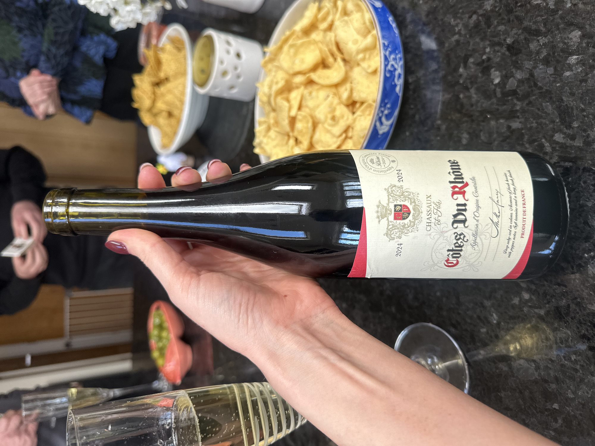 The image shows a person holding a bottle of red wine, specifically a "Côtes Du Rhône" from the producer "Chassaux et Fils," vintage 2024. The wine bottle is horizontal, held in the person's left hand, with a view of the wine label. Beside the hand and bottle is a champagne glass with visible bubbles. There are bowls of snacks such as chips in the background. A portion of a green dip appears behind the champagne glasses. Several other people are in the background. The surface is a dark countertop. The location and city of Tunbridge Wells, United Kingdom are not identifiable from the image.
