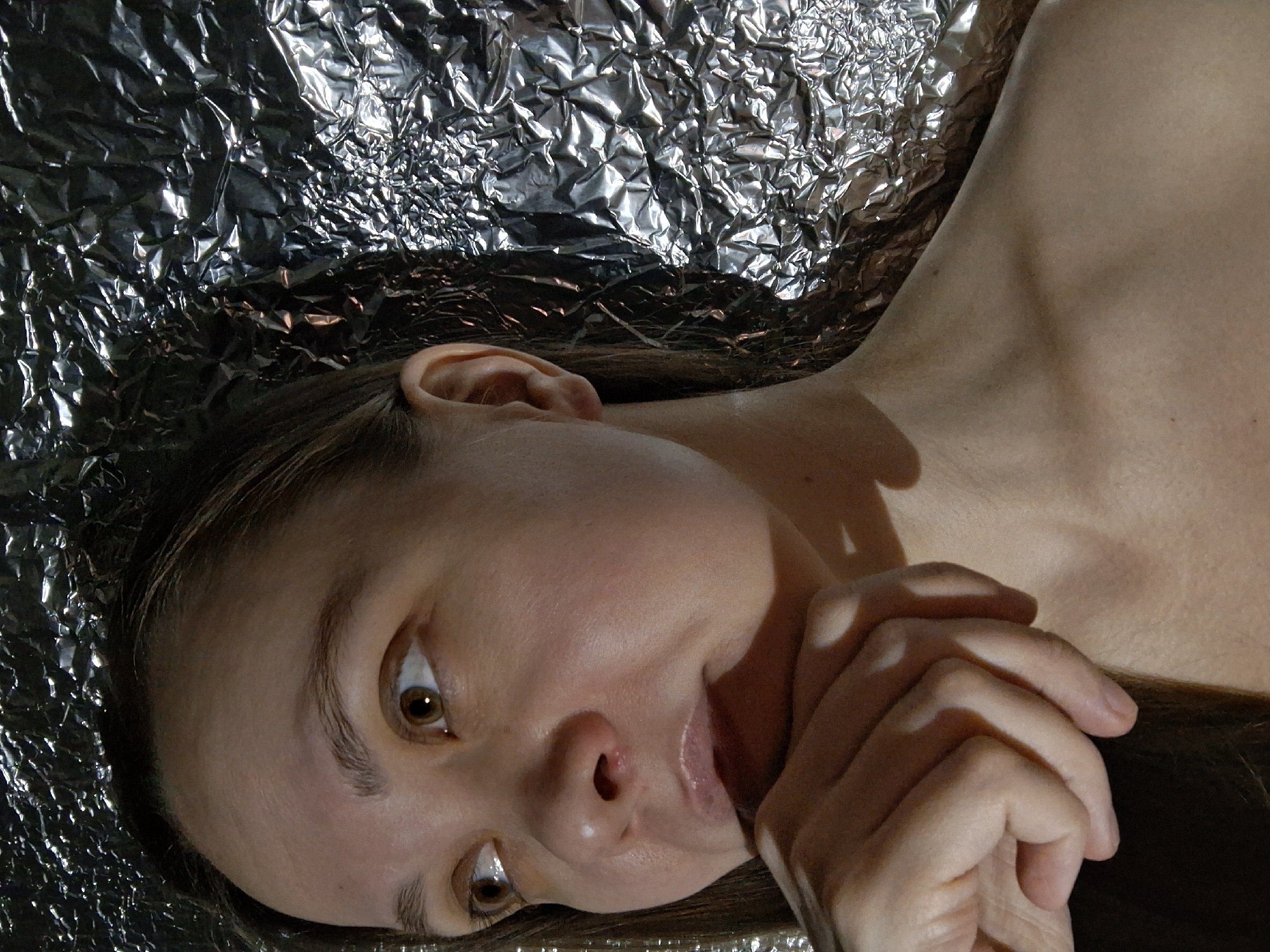 A person lies on a reflective, crumpled surface, possibly aluminum foil. The individual has fair skin, brown eyes, and light brown hair. Their face is positioned in the lower left of the frame, with their right hand near their chin. The background is composed of the crumpled, reflective material. There is no verifiable location in Chaikovsky, Russia.