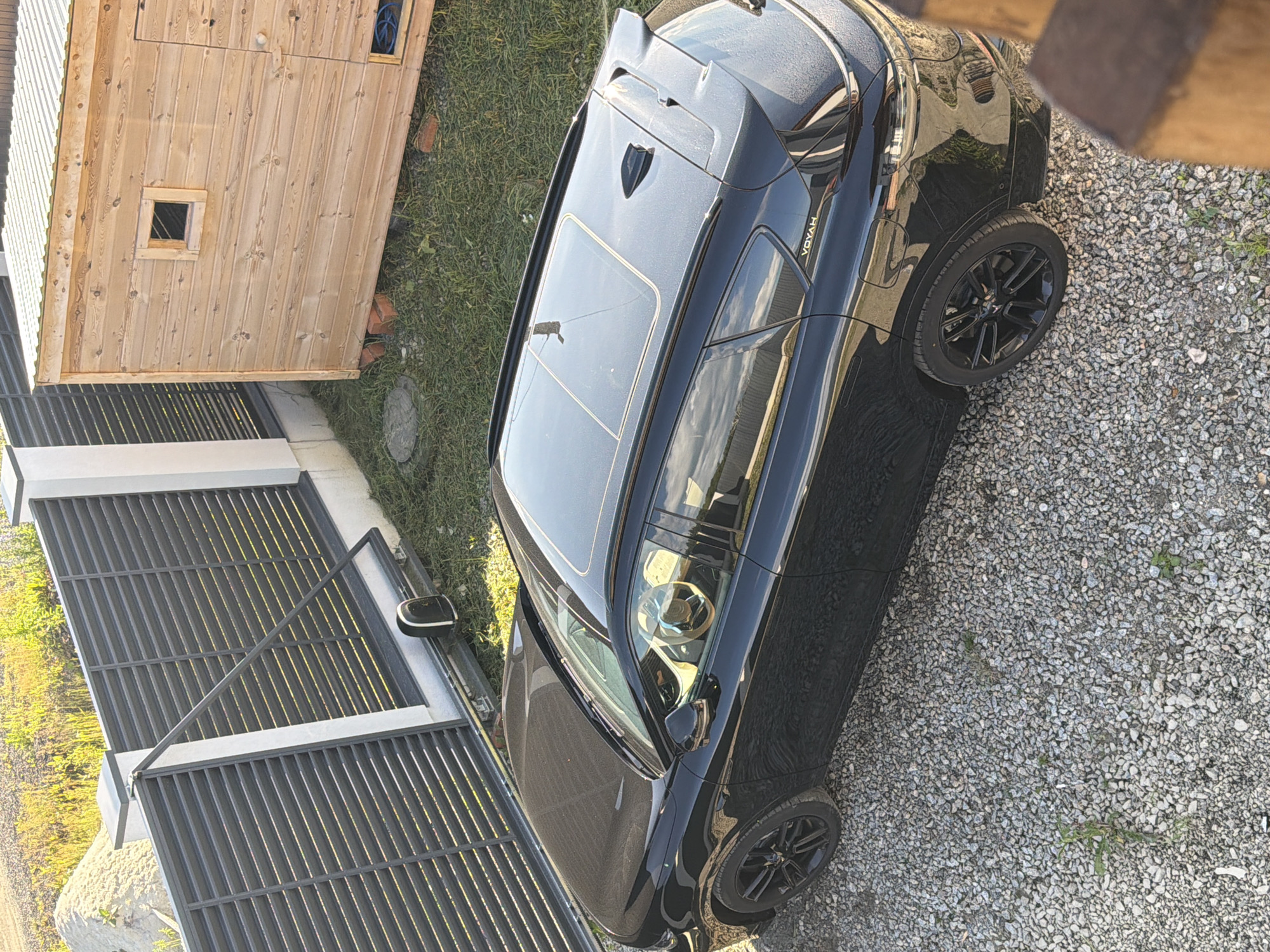 An overhead, eye-level shot captures a black Voyah SUV parked on a gravel surface. The vehicle, with its sunroof open, is positioned in front of a dark gray metal slat fence. To the right of the fence, a small wooden structure with a corrugated metal roof sits on a patch of green grass. The scene is illuminated by natural sunlight, casting subtle shadows. The background shows a gentle slope of dry vegetation. The image is likely taken in Malinovka, Russia.