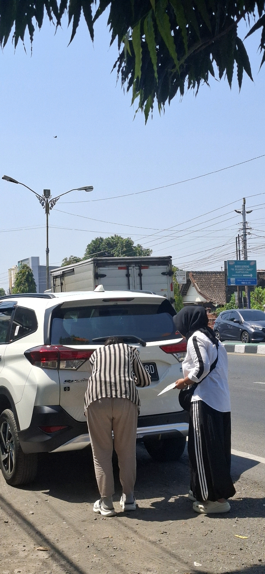 Two individuals are standing next to the open trunk of a white SUV on a street in Singopuran, Indonesia. The person closer to the car, wearing a black and white striped shirt and light brown pants, is leaning into the trunk, appearing to be retrieving or placing something. The second person, to the right and wearing a black hijab, white long-sleeved shirt with black stripes down the sleeves, and a black skirt with white stripes, holds a white piece of paper. The SUV has a license plate partially visible that reads "BD". In the background, a large truck is stopped, and further down the road, a dark-colored sedan is visible. The environment suggests a sunny day with a clear blue sky and some overhead power lines. A street light with decorative metalwork stands to the left, and a green directional sign is posted on the right side of the road. The ground is a mixture of dirt and gravel.