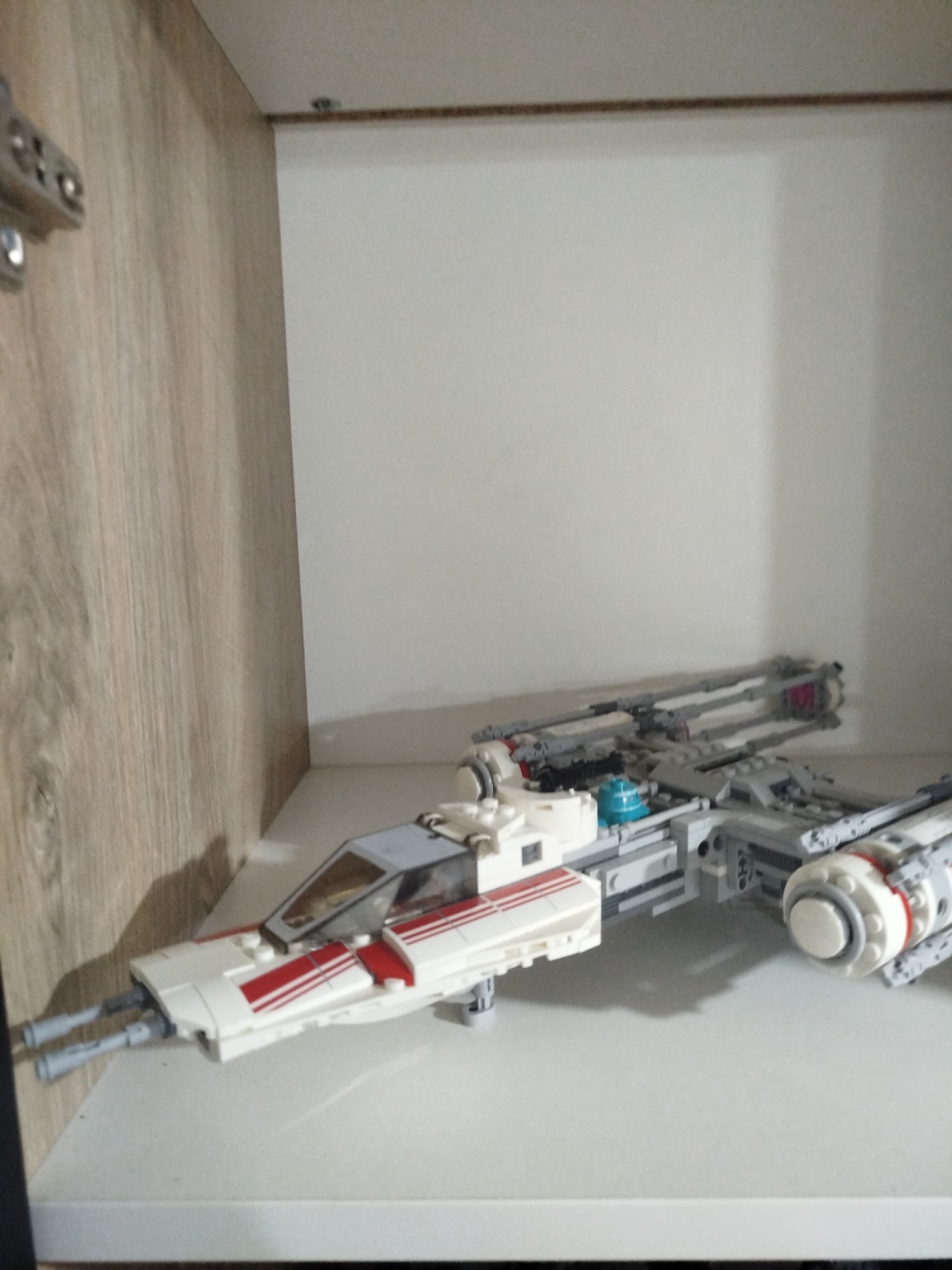 The image shows a Lego Y-wing starfighter model on a white shelf. The model is predominantly white with red stripes and grey accents. The starfighter is positioned on the right side of the frame, with its nose pointing towards the left. In the background, a plain white wall forms the backdrop, and to the left, a wooden-paneled surface is visible. The image is captured in a indoor setting, possibly a room or a display cabinet, and is not associated with the city of Mirny, Russia.