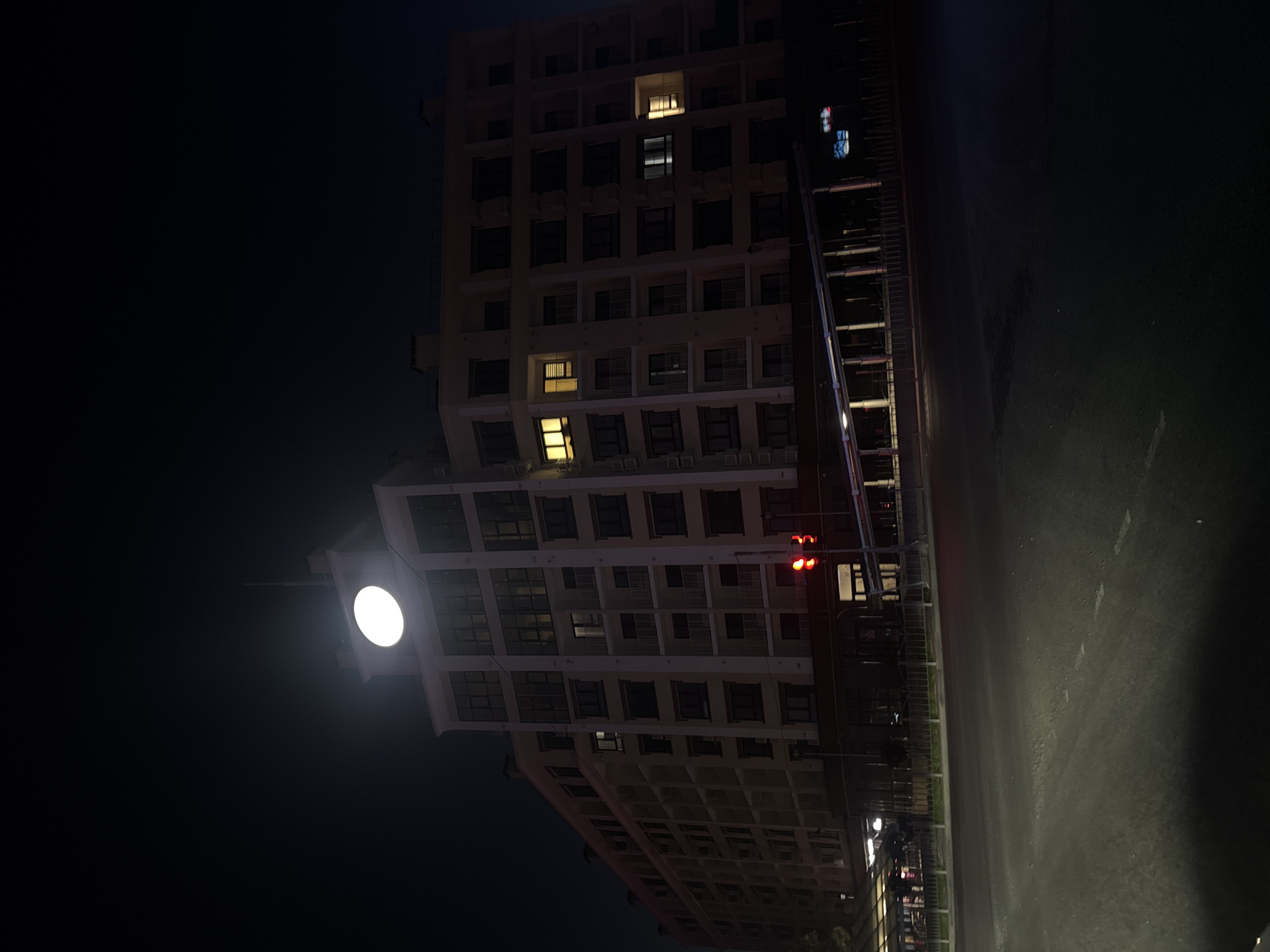This is a nighttime, low-angle, full shot of a large, multi-story building with a prominent clock face illuminated at the top. The building appears to be modern, with many windows and balconies. Some windows are lit, indicating occupancy. In the foreground, there is a street with a traffic light showing red. The scene is set in the city of Mariupol, Ukraine.