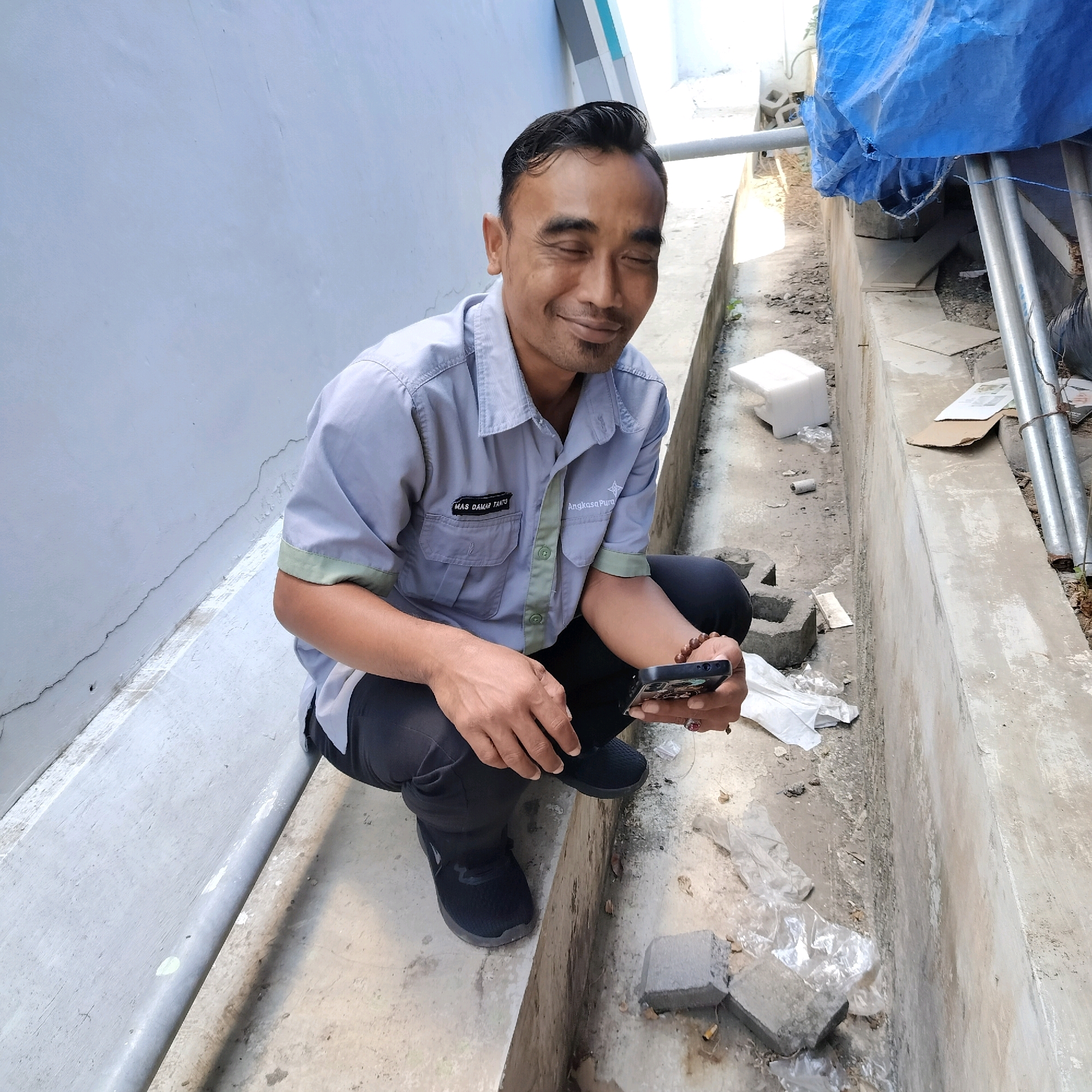 In Sleman, Indonesia, a man wearing a light grey short-sleeved collared shirt with green trim and black pants kneels beside a concrete drainage channel. His eyes are closed, and he is smiling as he holds a dark-colored smartphone in his right hand. The shirt has two patches: one on the left chest pocket reads "MAS DAMAR TAMTU," and a larger logo with star-like elements and text "Angkasa Pura" is on the right chest. He wears a beaded bracelet on his right wrist and a ring on his right index finger. The man is positioned in an outdoor environment with a light blue wall to his left and the drainage channel to his right. The channel contains debris, including plastic sheeting and construction materials. In the background, a blue tarp is draped over metal pipes, and a white styrofoam block is visible. The ground is concrete and dirt.