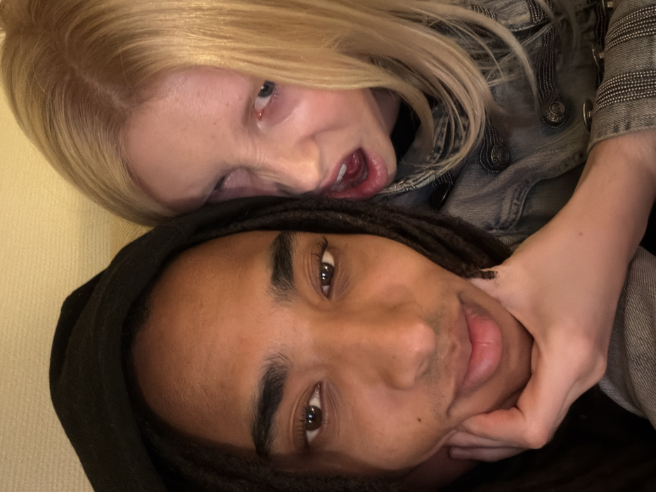 The image displays a close-up indoor portrait of two individuals. The precise location within the city of Paris, France, is not visually discernible from the image, which features an indistinct light-colored background.
On the left, a person with dark skin and long dark dreadlocks, wearing a dark hooded garment, occupies the majority of the left frame. This individual has dark brown eyes and is looking directly at the camera with a neutral expression. Their chin rests upon their right hand, with fingers curled beneath the jawline. A faint stubble is visible on their chin and upper lip.
To the right and slightly behind the first individual, a person with fair skin and long blonde hair is partially visible. This individual is wearing a light-colored denim jacket with prominent silver buttons. They are making an exaggerated facial expression, characterized by furrowed brows, a wrinkled nose, bared teeth, and an open mouth. Their left eye is slightly narrowed, and their right eye is partially obscured by hair. Their left hand is positioned on the chin and lower jaw area of the first individual, appearing to grip or cup it. The interaction between the two suggests a playful or theatrical pose.