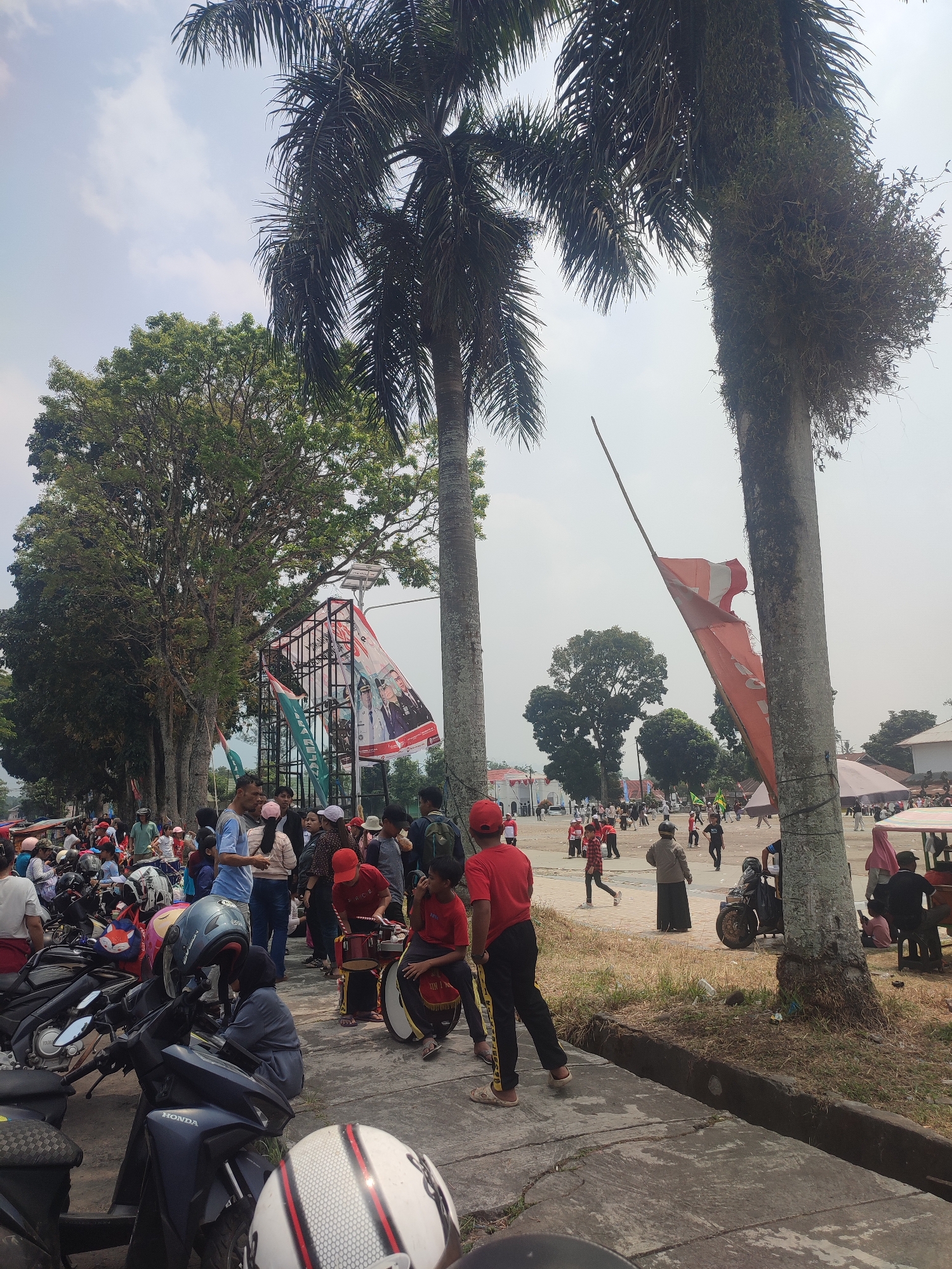 The image depicts a street scene in Dwi Tunggal, Indonesia, characterized by a gathering of people on a paved area adjacent to a grassy embankment. Two prominent palm trees dominate the foreground, with a large, mature deciduous tree on the left side of the frame. In the background, a temporary stage structure draped with banners featuring portraits of individuals is visible. Several individuals, including children and adults, are present, with some appearing to be engaged in a drum performance. Motorcycles are parked along the left side of the scene. An orange flag is prominently displayed on a pole, leaning significantly to the right. The sky is overcast with scattered clouds. The environment suggests an outdoor public event or gathering.