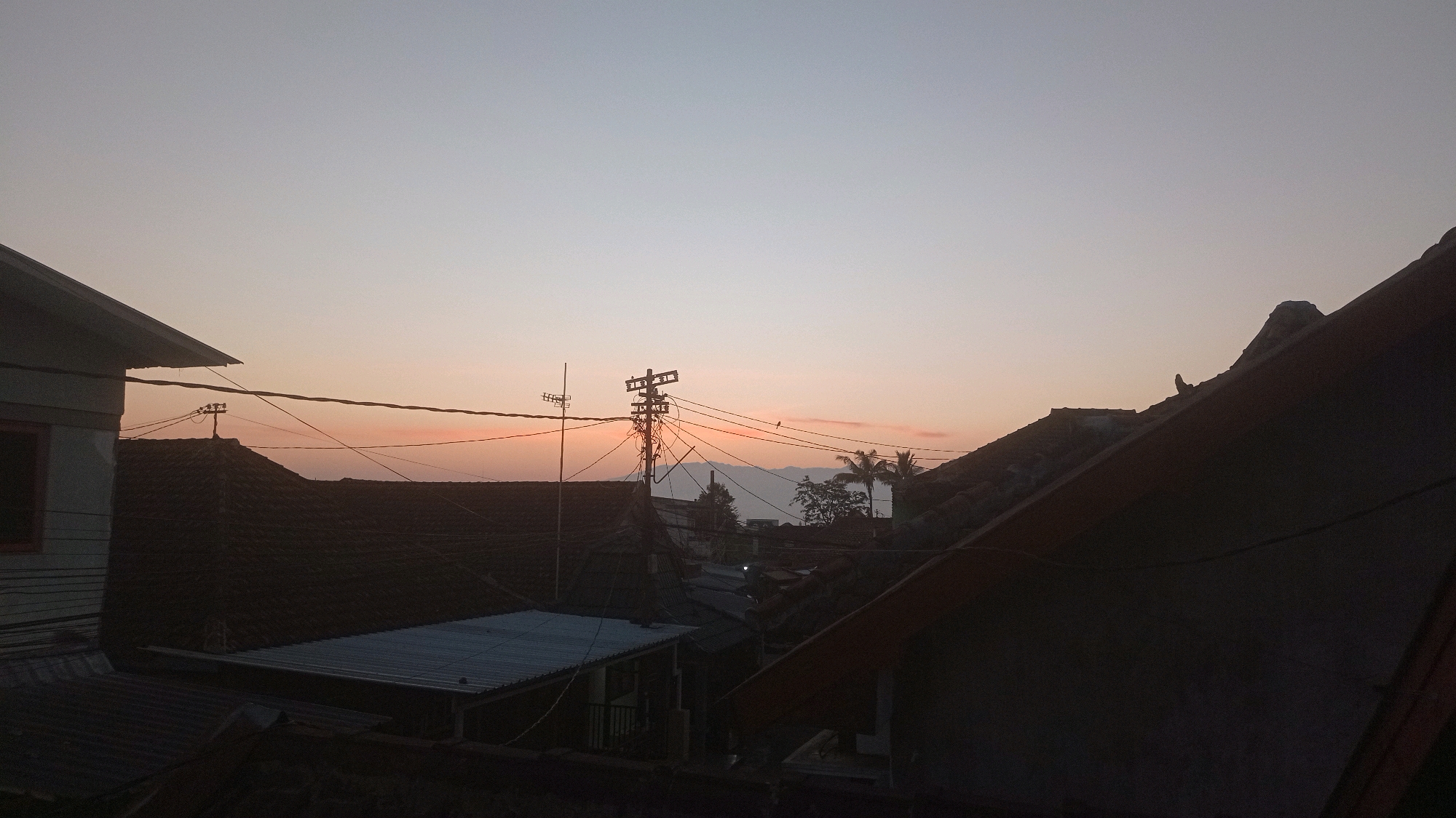 The image captures a twilight scene over a residential area in Mulyoagung, Indonesia, likely from an elevated vantage point such as a rooftop. The sky exhibits a gradient of soft, muted colors, transitioning from a pale blue at the top to a warm, peachy hue near the horizon, suggesting either sunrise or sunset. Silhouetted against this sky are the rooftops of numerous houses, characterized by their dark, tiled surfaces.
Prominently featured is a utility pole with a complex network of wires crisscrossing the frame. The pole itself appears to be made of metal and is equipped with insulators and other electrical equipment. In the distance, the faint outlines of mountains are visible. Several trees, including what appear to be palm trees, are also present, adding to the tropical feel of the environment. The foreground is dominated by the dark, angular shapes of more rooftops and structural elements, possibly belonging to the observer's own dwelling. No people are clearly discernible in the image, and there are no explicit actions or interactions depicted. The overall impression is one of a quiet, domestic landscape bathed in the soft light of dusk or dawn.