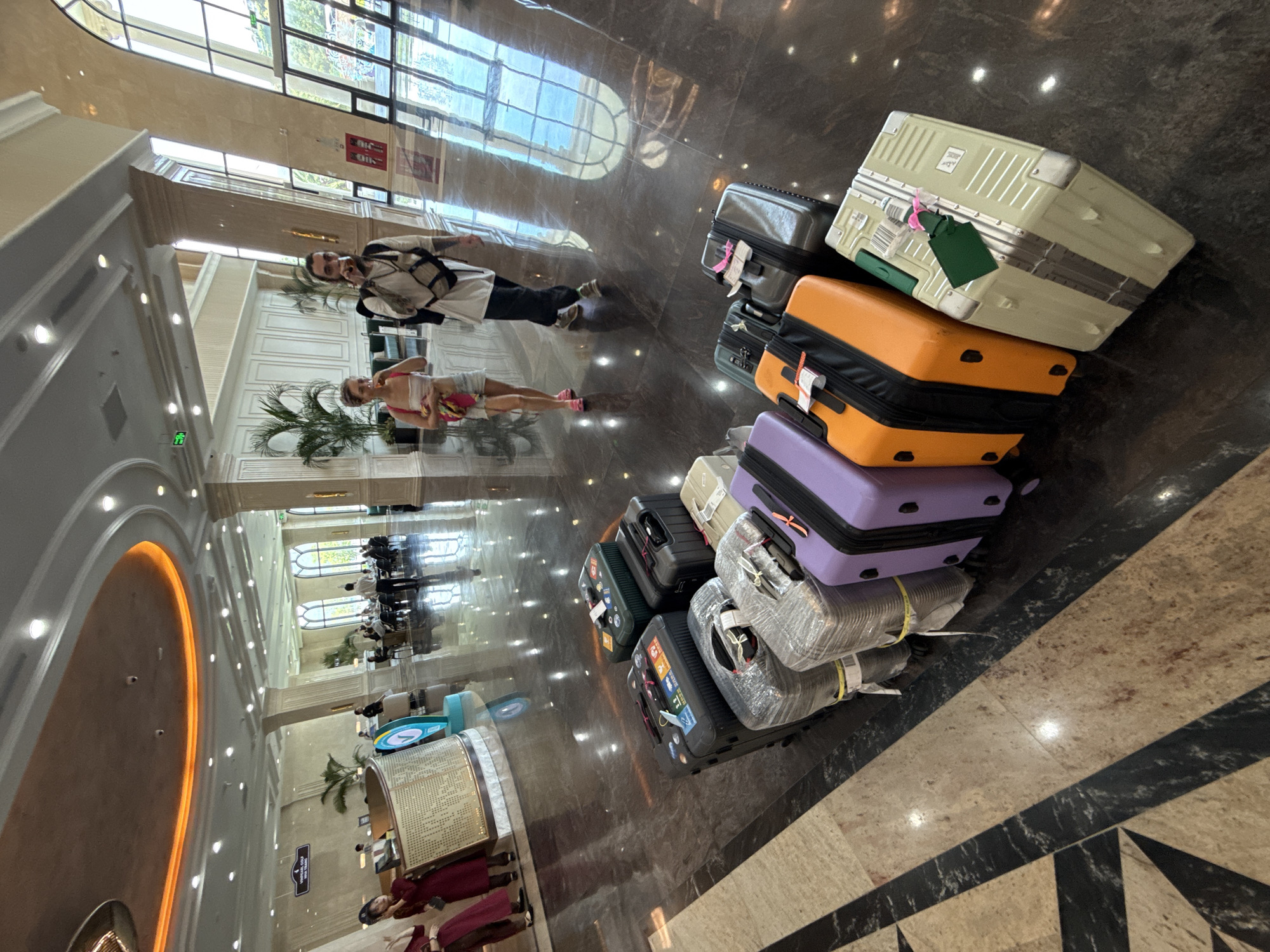 The photograph depicts the lobby of a hotel in Nha Trang Ward, Vietnam. The floor is shiny, reflective marble with dark and light brown tones in a patterned design. A stack of approximately 8-10 suitcases of varying colors (including beige, silver, orange, purple, green, and black) sits on the right side of the frame. The suitcases are stacked haphazardly.
A couple stands near the center of the frame. The man is wearing a white shirt and black pants, while the woman is in a lighter colored dress or top and shorts. They are positioned as if they were standing or walking, but their reflection in the shiny floor makes it appear as though they are lying down.
In the background, the hotel lobby stretches out, featuring high ceilings, decorative columns, and a reception desk. A group of people in reddish-burgundy uniforms can be seen near the reception area. Natural light streams in through large windows. The scene appears to be a typical check-in/check-out area in a hotel.
