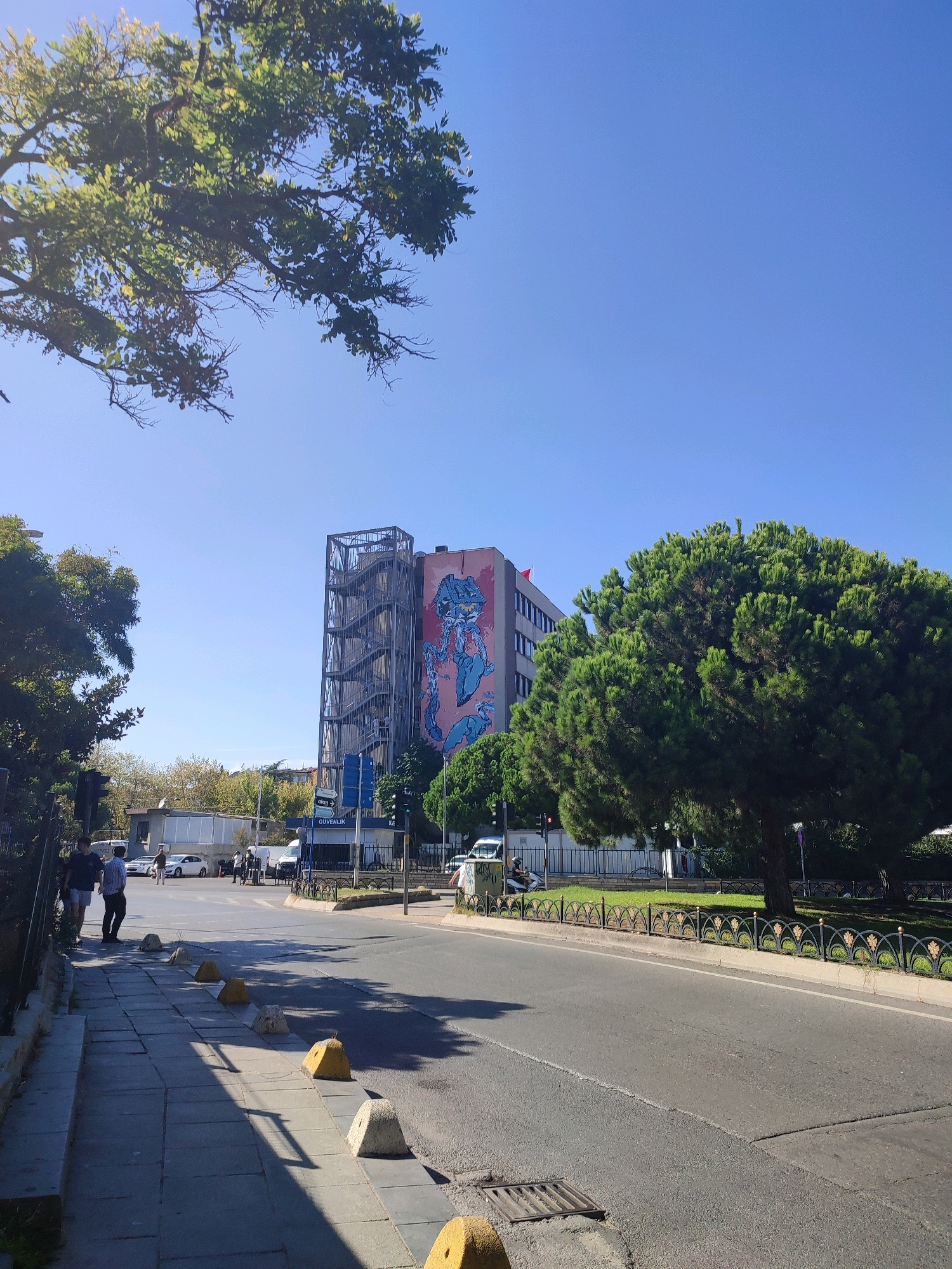 The image depicts a street scene in Kadıköy, Turkey, on a clear, sunny day. A sidewalk lined with concrete bollards runs along the left side of a paved road. Several people are walking on the sidewalk. To the right of the road is a green grassy area with trees and an ornate metal fence. In the background, a multi-story building is visible, featuring a large mural of a blue creature and an external metal fire escape. Cars and a van are parked or driving on the road. A traffic light is visible. The sky is a clear, bright blue.