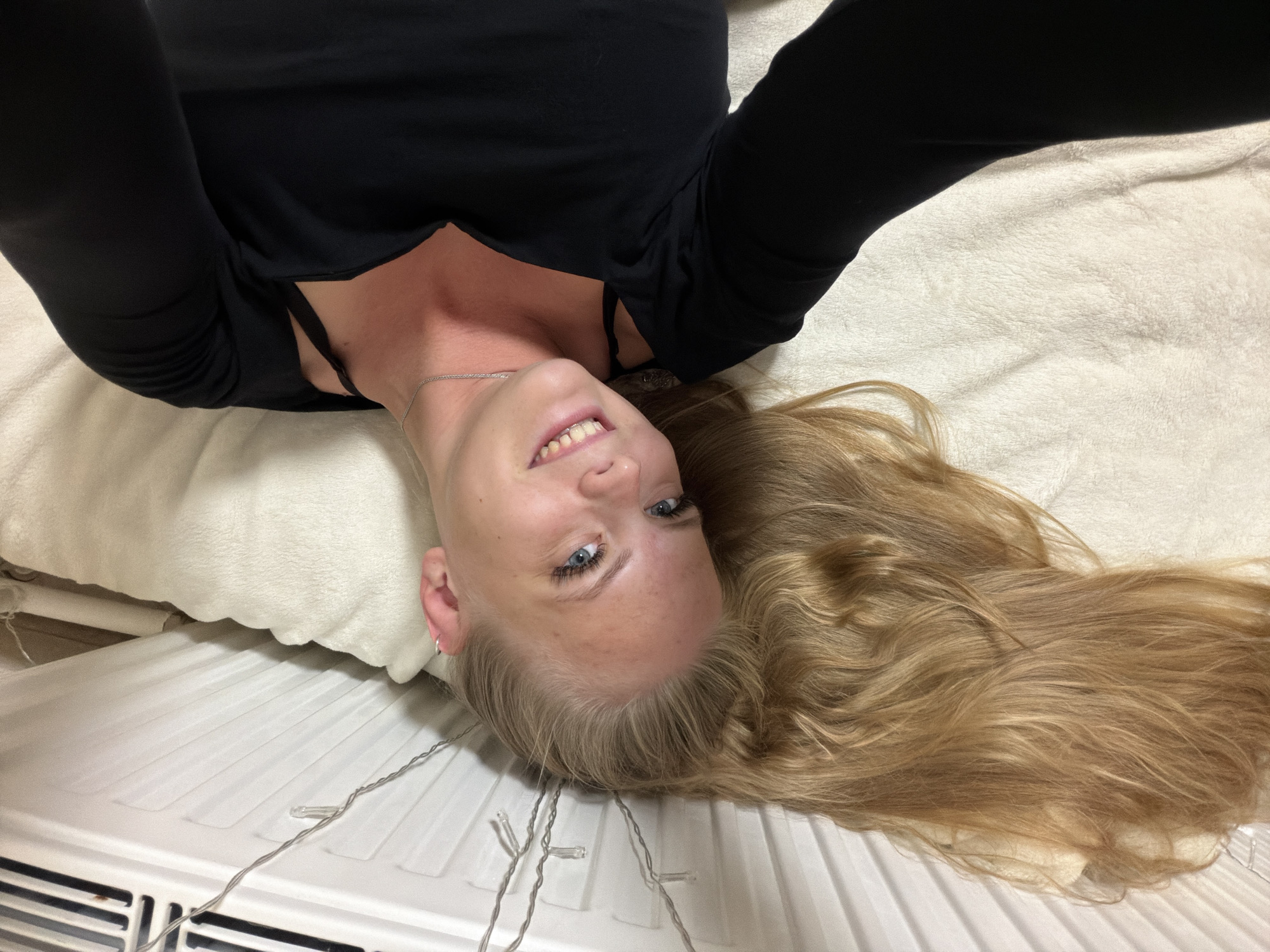 The media file is a close-up, upside-down selfie of a young woman lying on a white, plush surface, which appears to be a bed or sofa. She has long, blonde hair that is spread out around her head. Her head is resting on a white pillow. She is wearing a black long-sleeved top and a black bra. A thin silver necklace is visible around her neck. Her eyes are blue, and she is smiling. She is also wearing a small silver hoop earring in her visible ear. In the background, a white radiator with vertical slats is partially visible, with some string lights draped across it. The lighting suggests it is likely indoors, possibly in the evening or a dimly lit room. The overall mood is relaxed and playful. There is no visible text in the image.