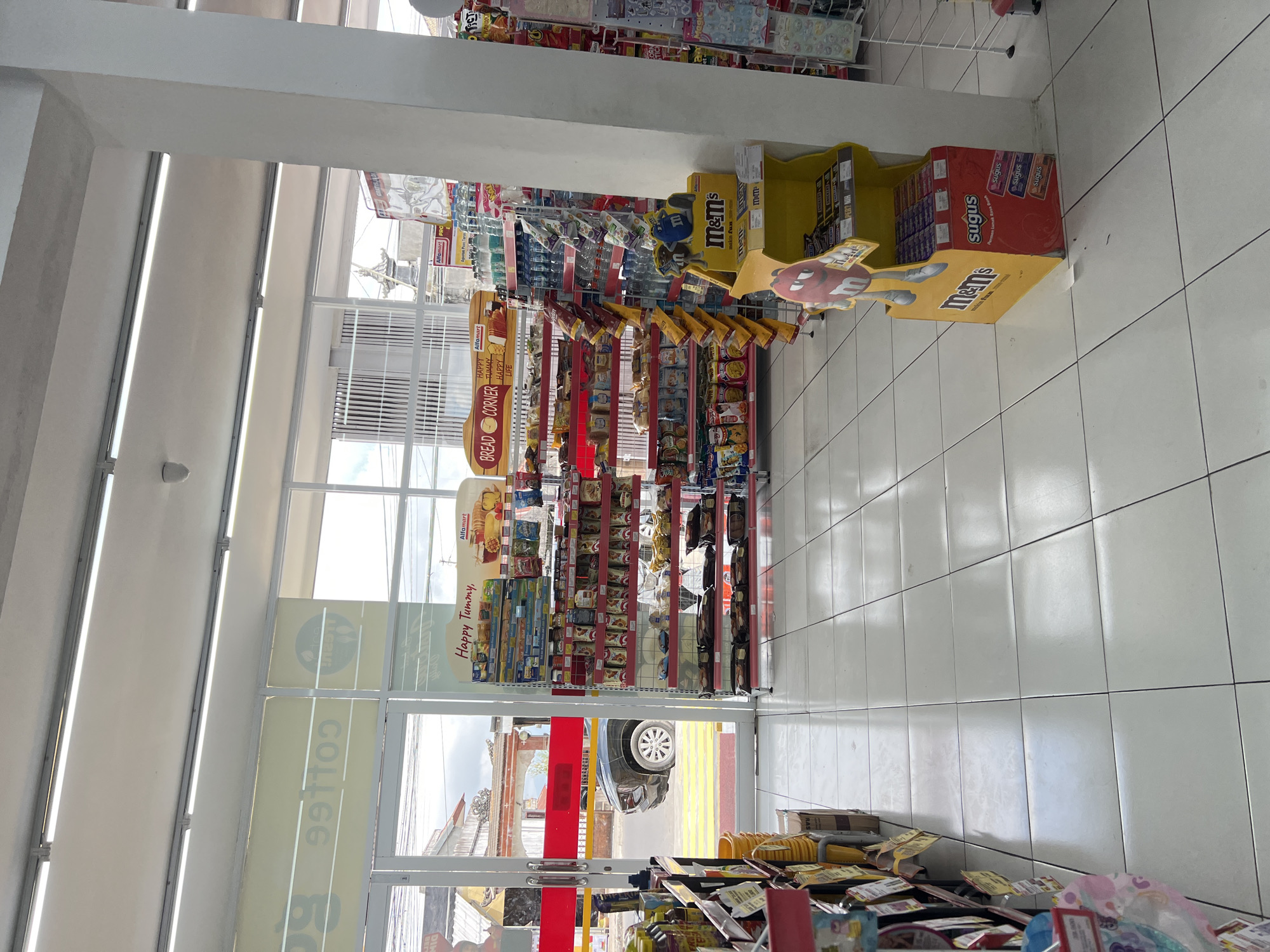 The image depicts the interior of an Alfamart convenience store in Kintamani, Indonesia. The store features bright fluorescent lighting and a polished white tiled floor. Prominently displayed are shelves stocked with various packaged goods, including snacks, drinks, and candy. To the right, a large yellow display stand for M&M's candy is visible, with additional Sugus candy packets below. Near the entrance, a rack of bottled water and other beverages is organized. Through the glass doors, a black car is parked outside, with a glimpse of a street and buildings. The overall scene suggests a well-maintained and typical convenience store environment.